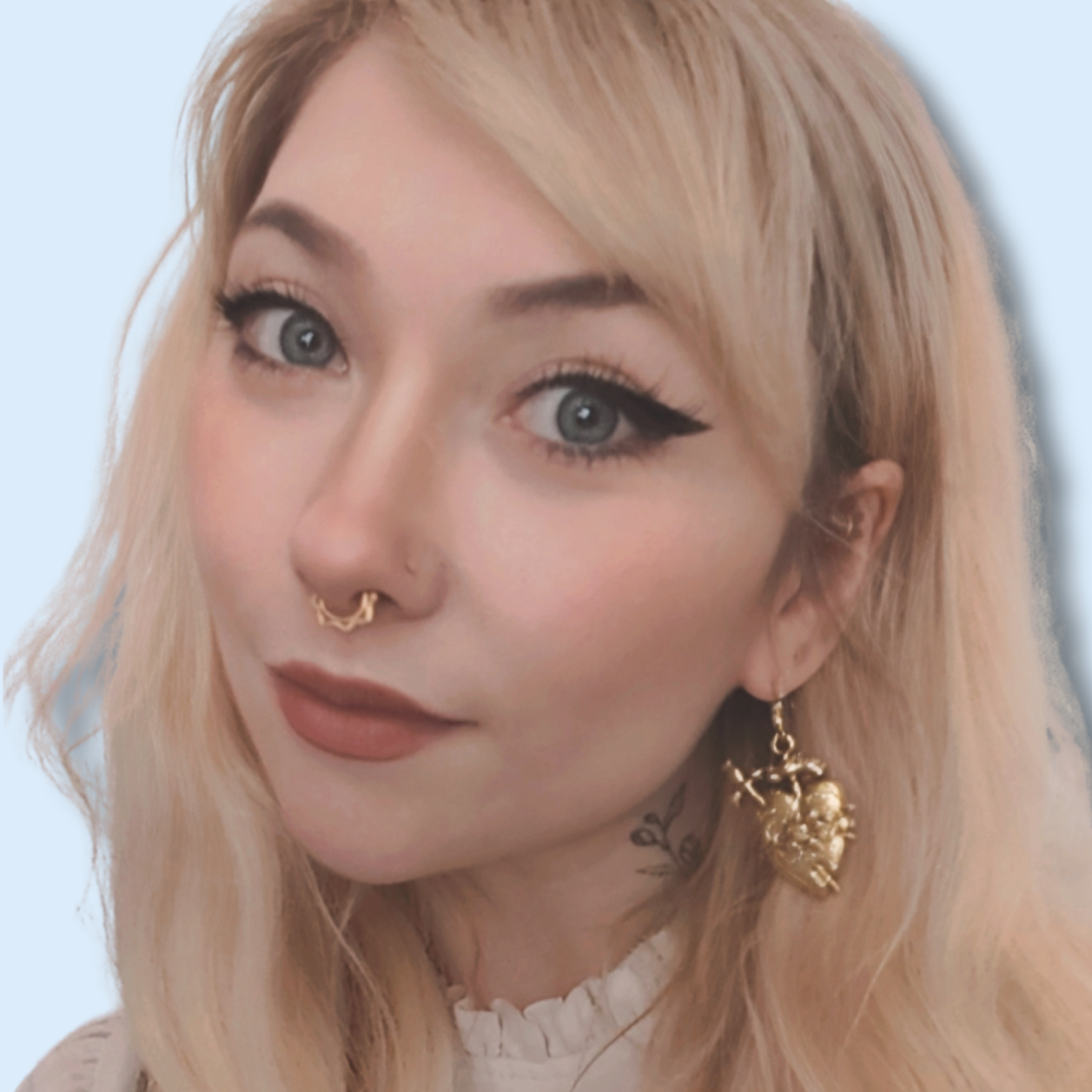 Close-up of a woman with blonde hair, blue eyes, winged eyeliner, and gold jewelry, including a nose ring, a heart-shaped earring, and a tattoo on her neck.