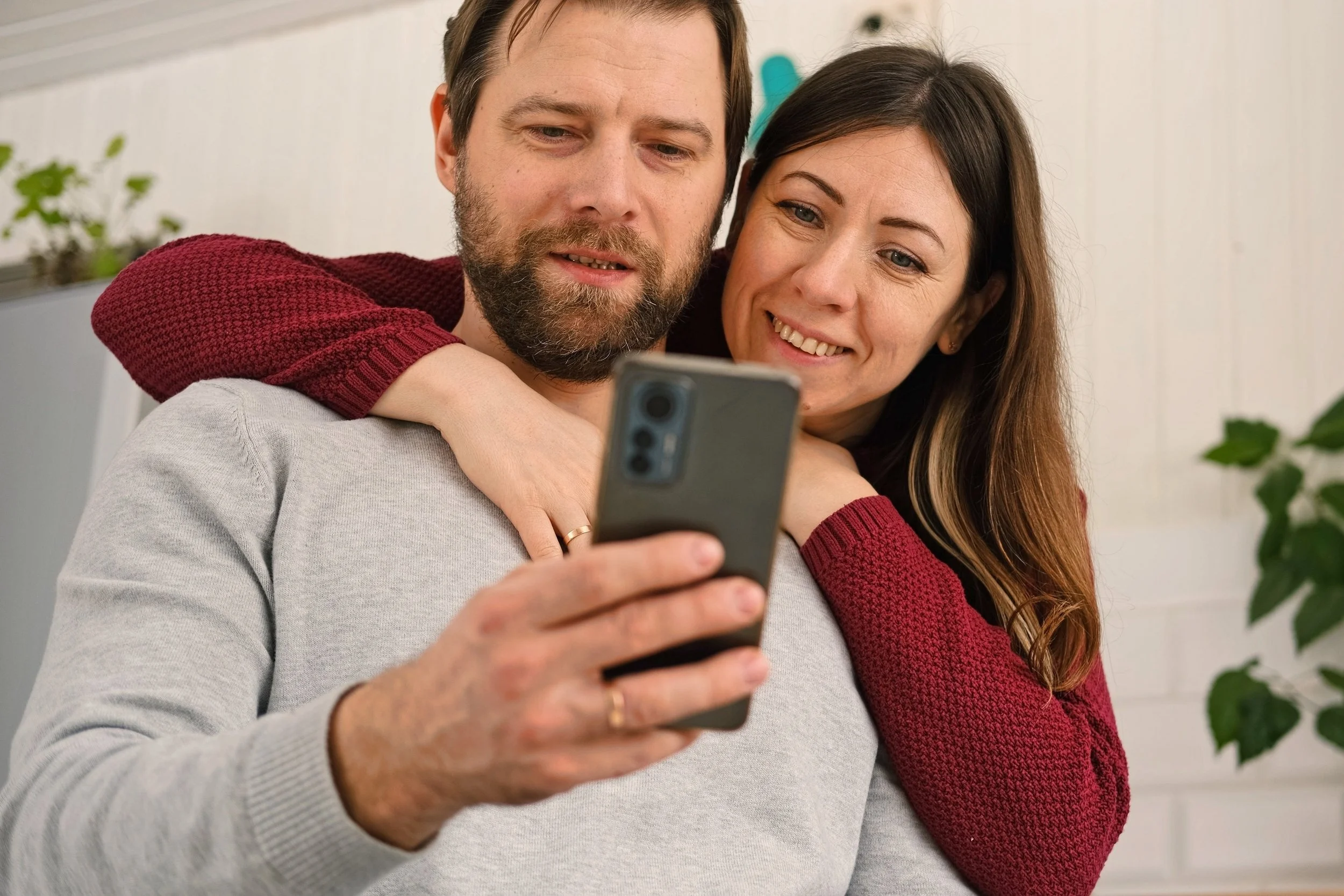 a man wearing a grey sweater while holding a dark-colored smartphone, with a woman in a red sweater smiling as she embraces him from behind.