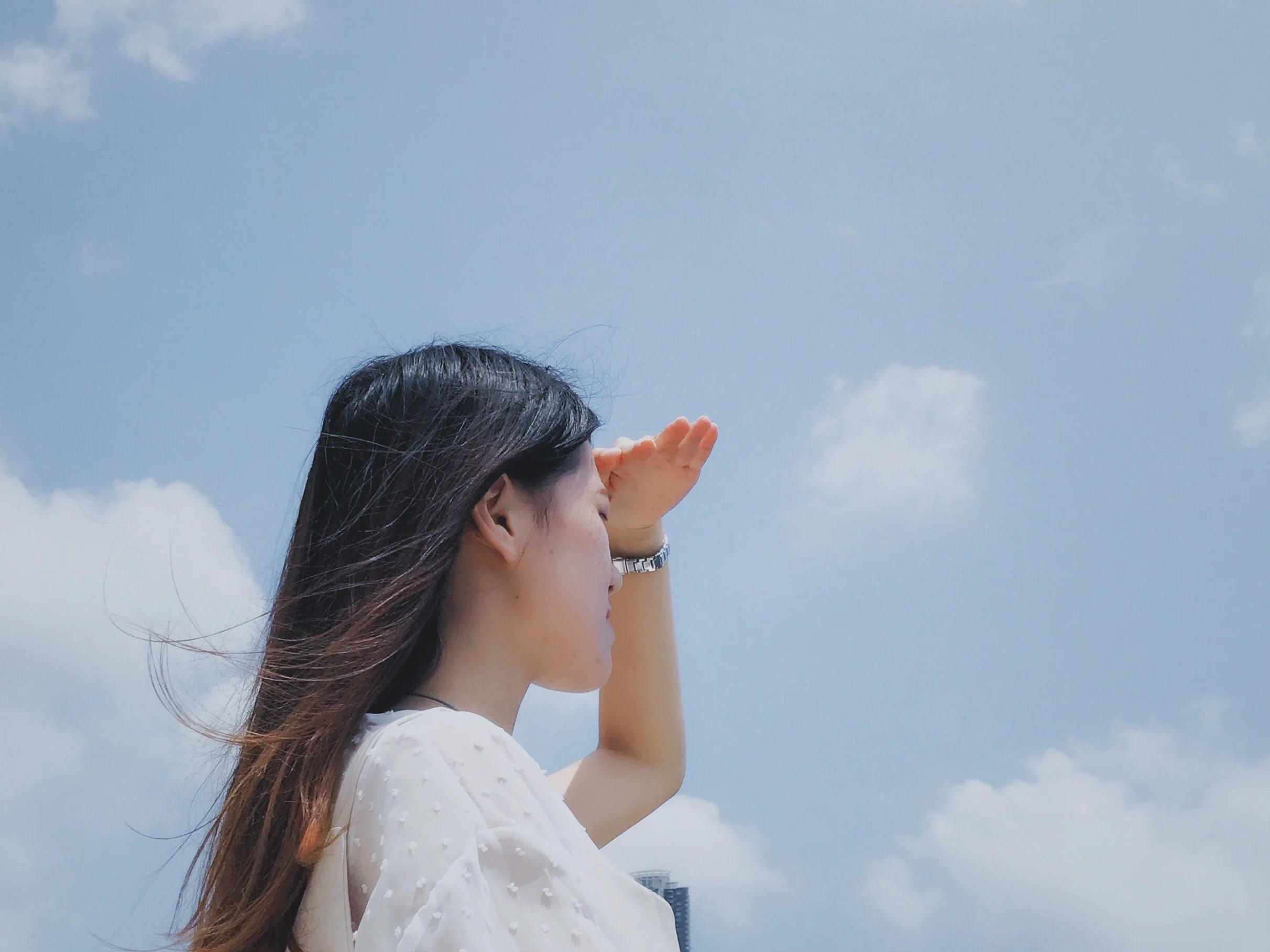 A woman with long hair wearing glasses and a white dress, standing outdoors on a sunny day with a clear blue sky and some clouds.