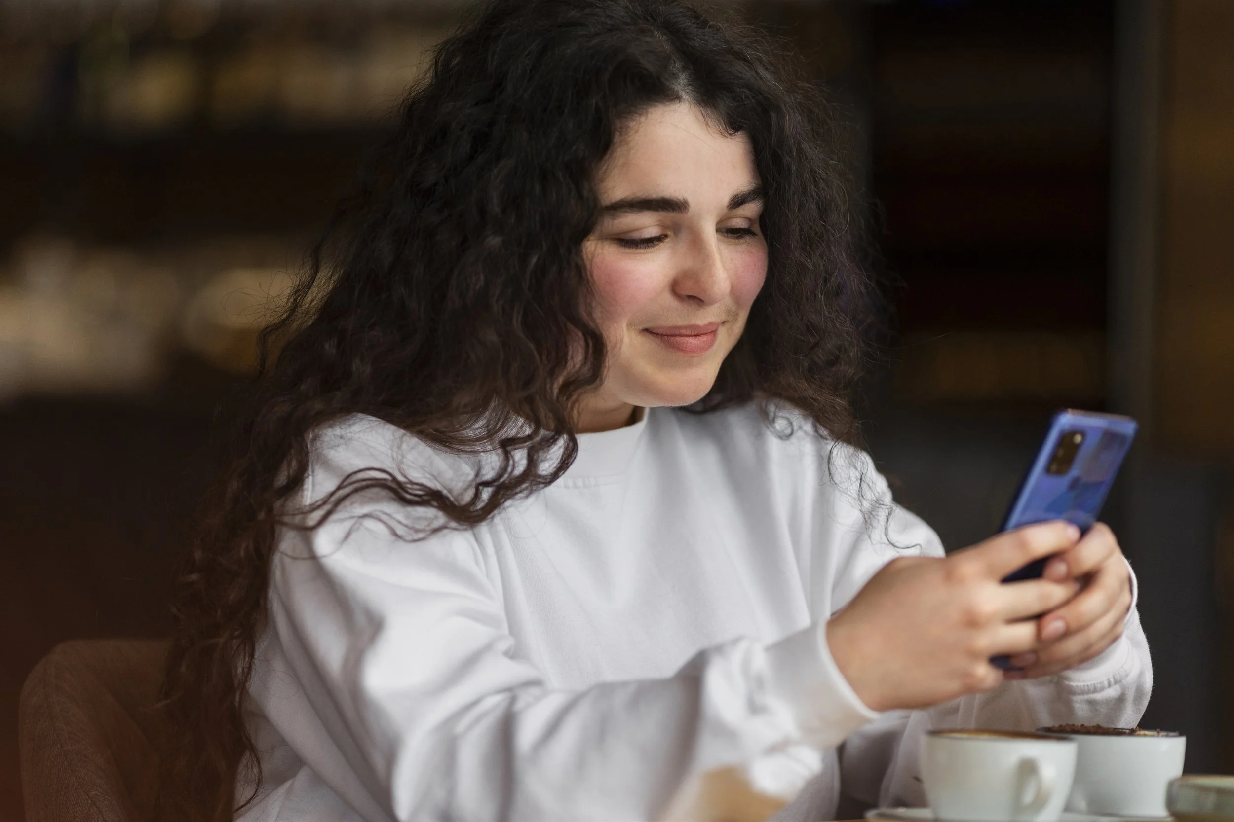 A young woman with long, curly dark hair is smiling while looking at her phone, sitting at a table with a bowl of food in front of her.