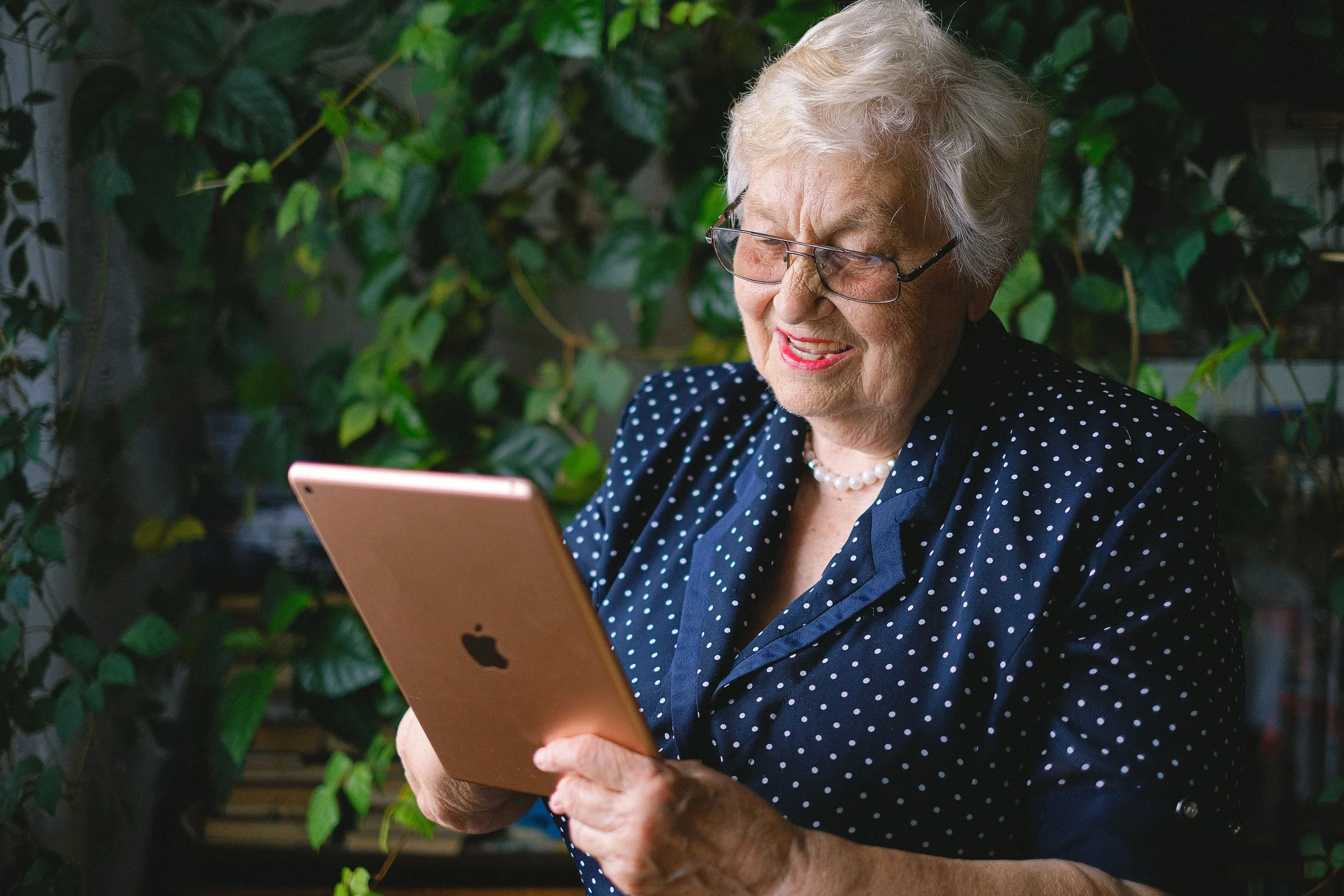 An older woman is sat smiling and looking at an iPad.