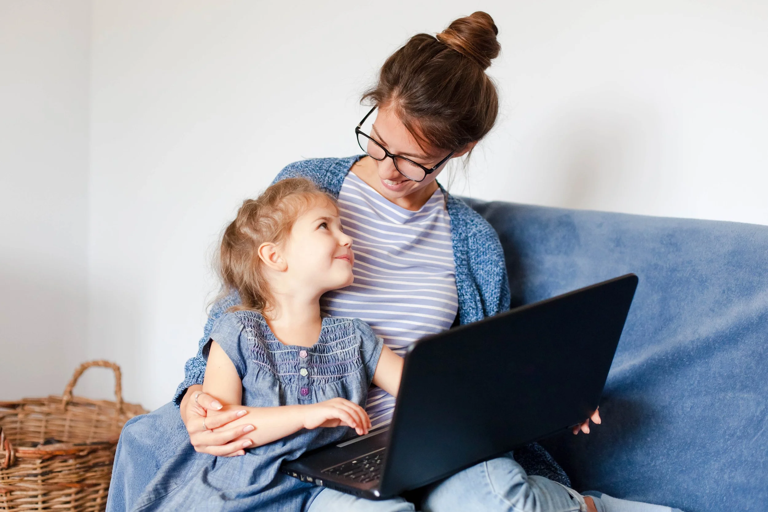 A woman with glasses and a bun hairstyle sitting on a blue couch with a young girl, both looking at a laptop screen. The woman is smiling at the girl, who is looking up at her.