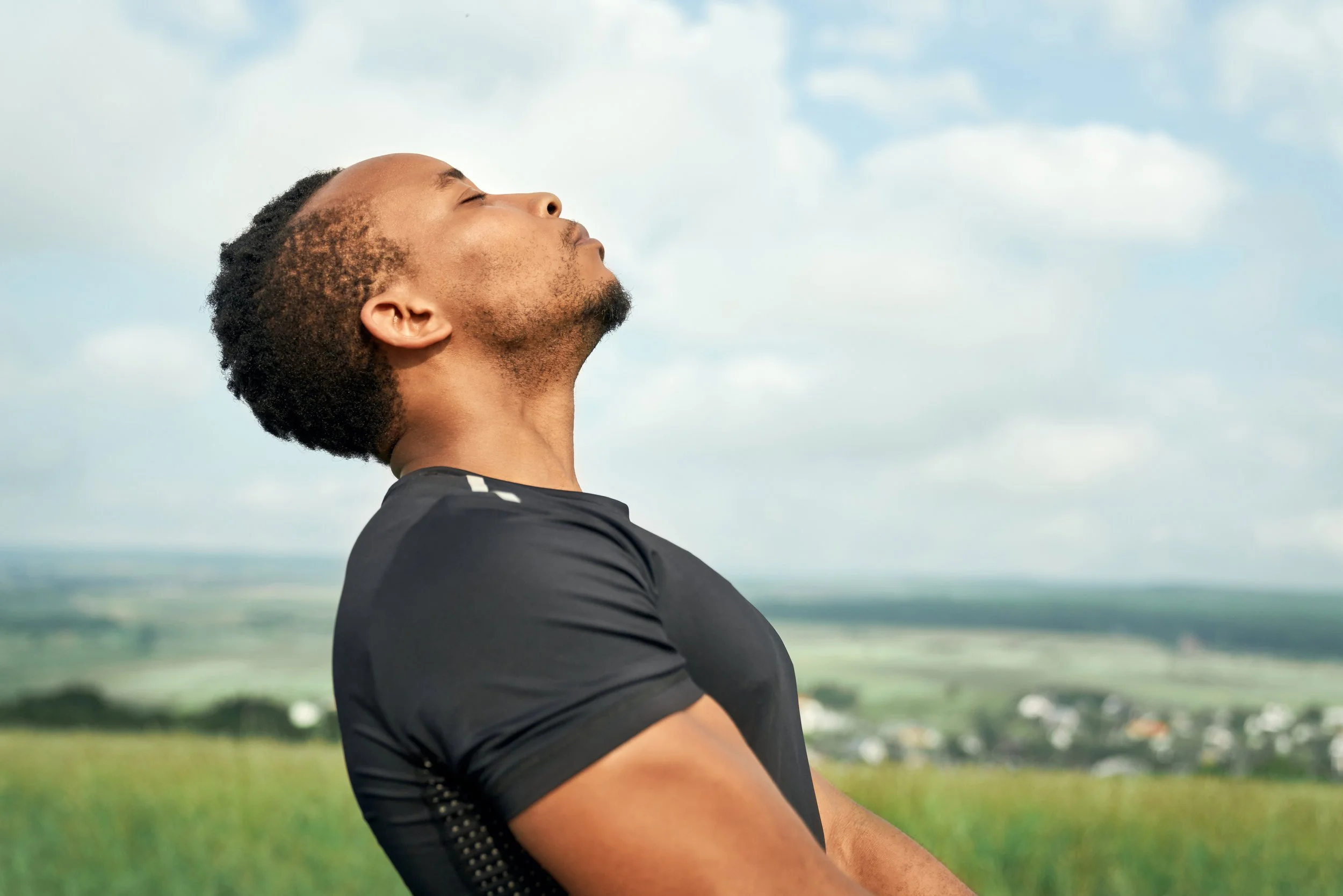 A man with closed eyes and relaxed expression standing outdoors against a cloudy sky, wearing a black sports shirt.