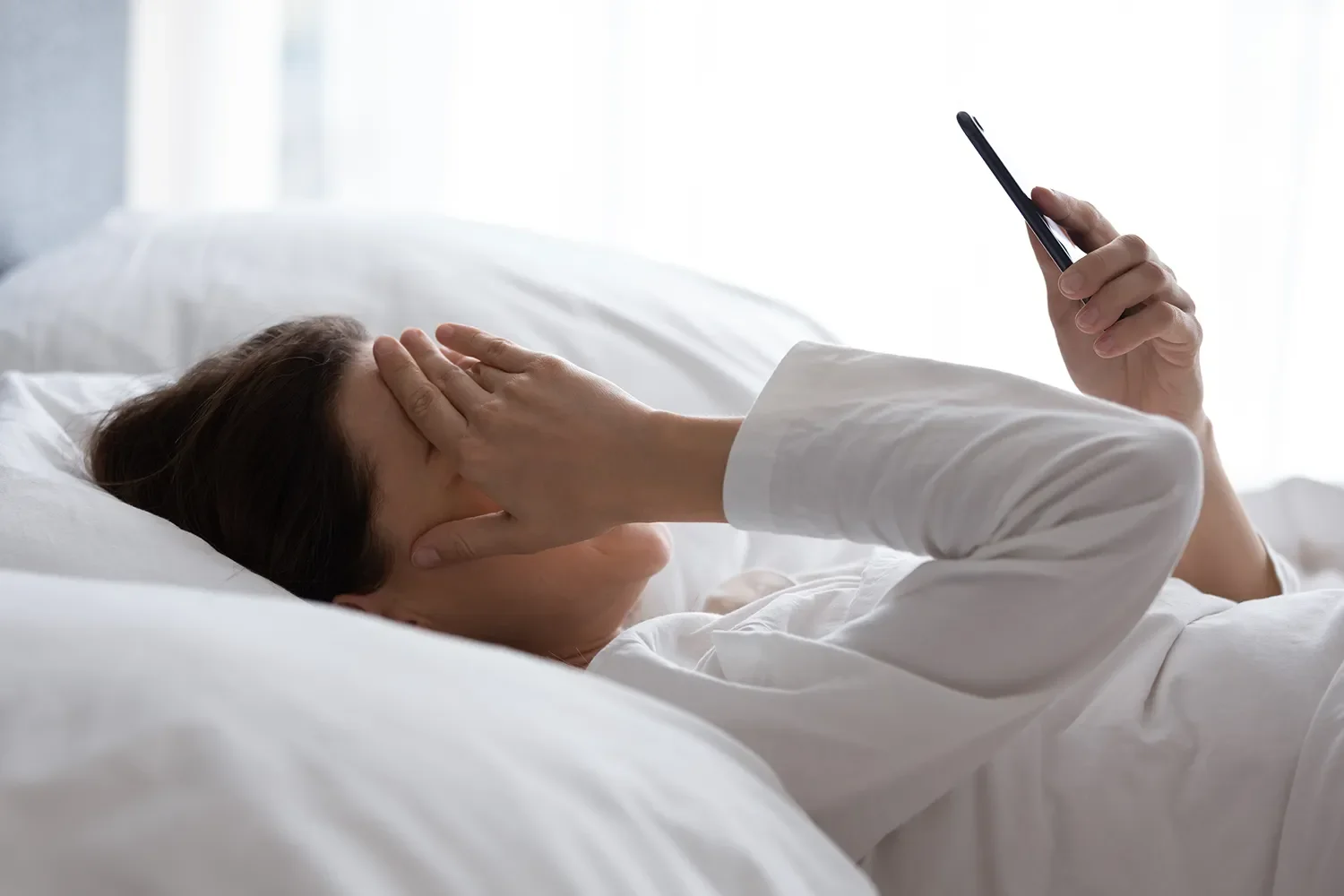 Person lying in bed, holding forehead with one hand, looking at smartphone with the other hand.