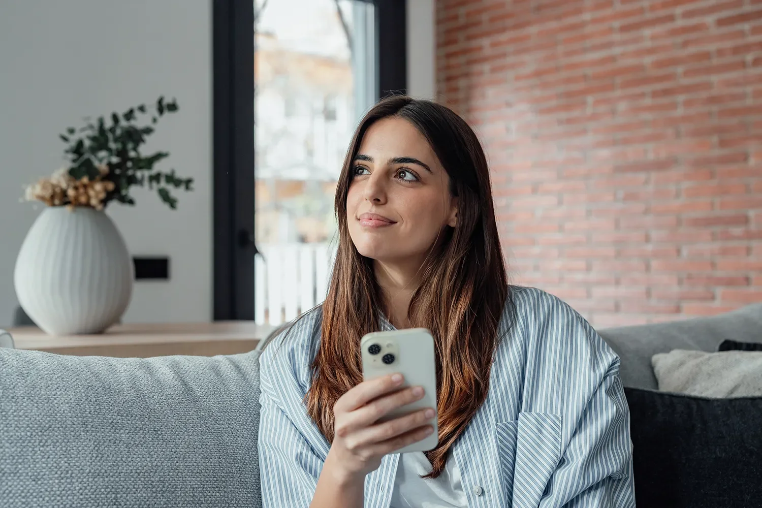 A woman sitting on a gray couch, holding a smartphone, smiling and looking up to her right inside a modern room with a brick wall and a large window.