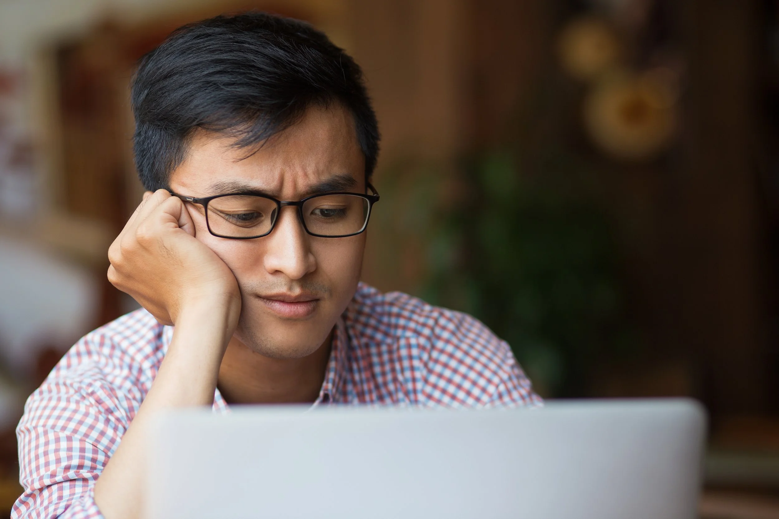 Young man with black hair, glasses, and a checked shirt looking at a laptop screen with a concerned or focused expression.