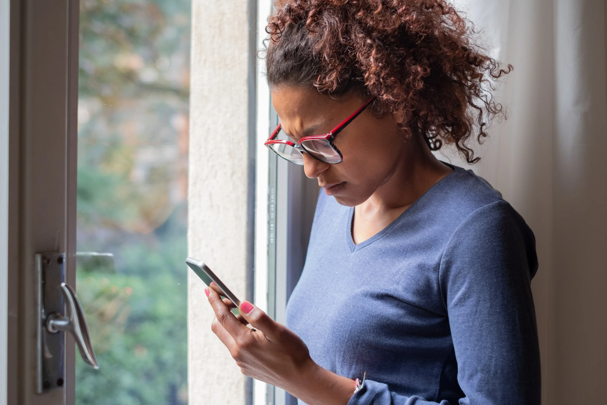 Woman with curly hair and glasses looking down at her phone while standing by a window.