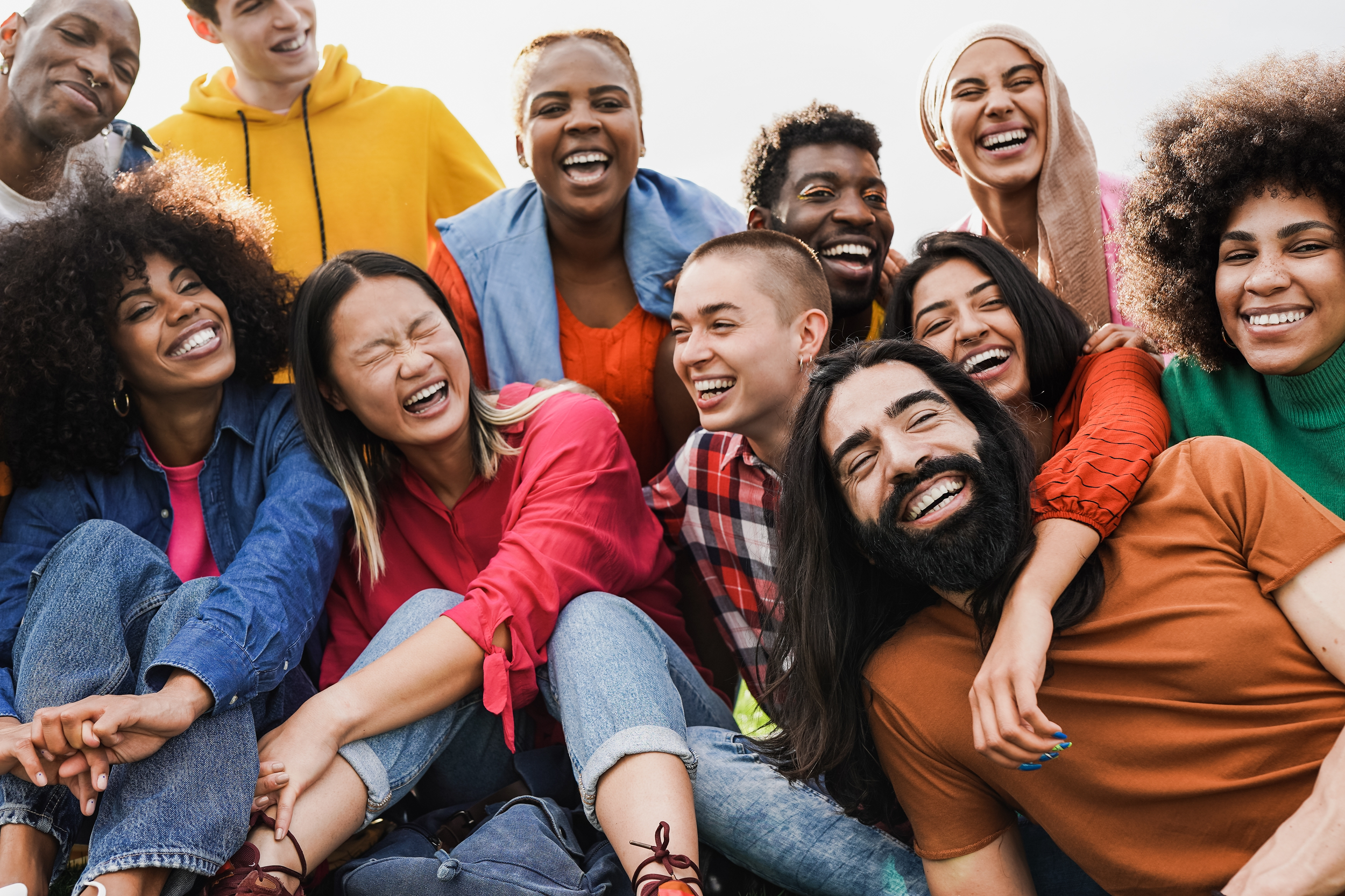 A diverse group of people laughing and smiling, sitting closely together outdoors, enjoying each other's company.