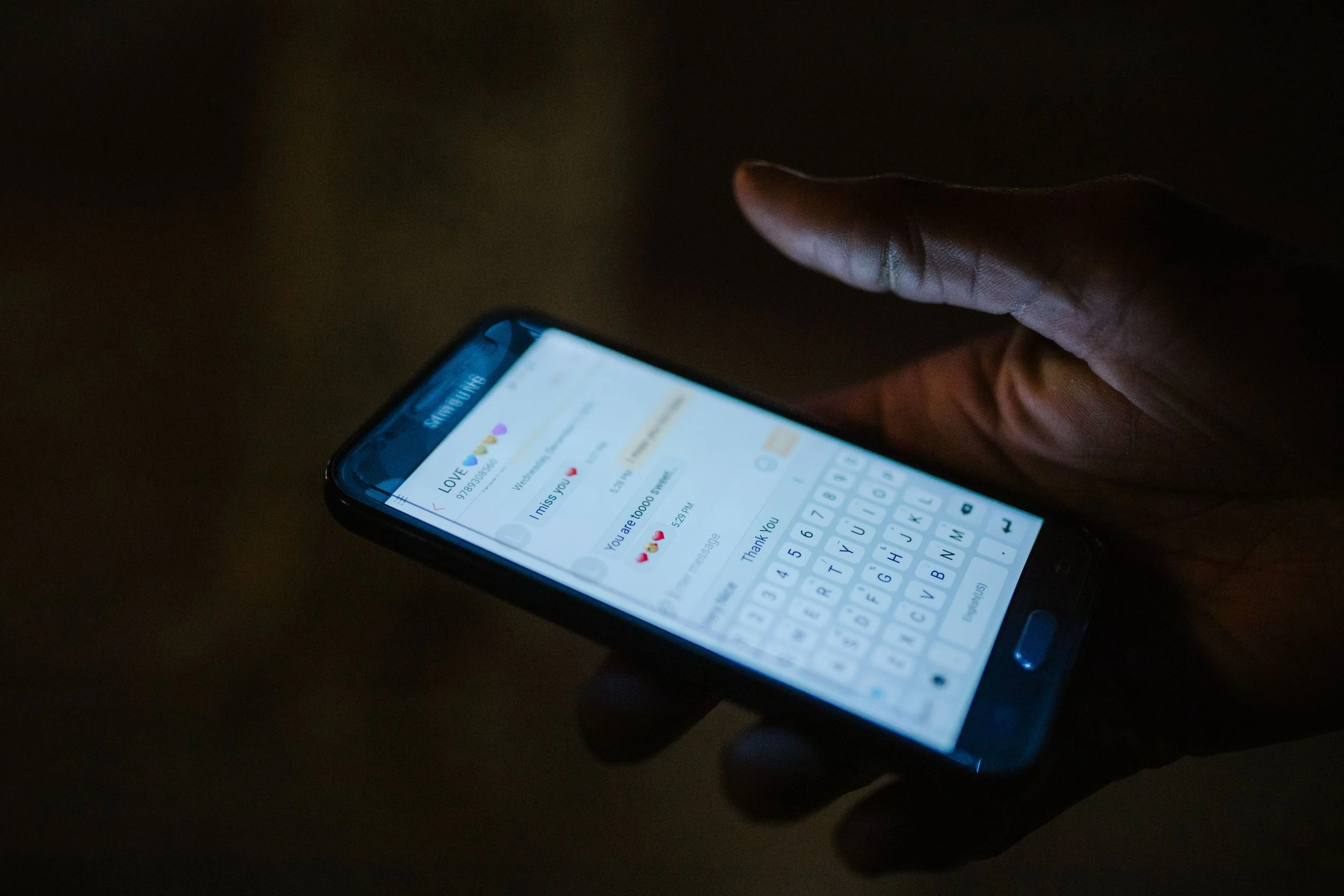 A hand holds a phone in the dark, showing a romantic messaging conversation with heart emojis on screen.