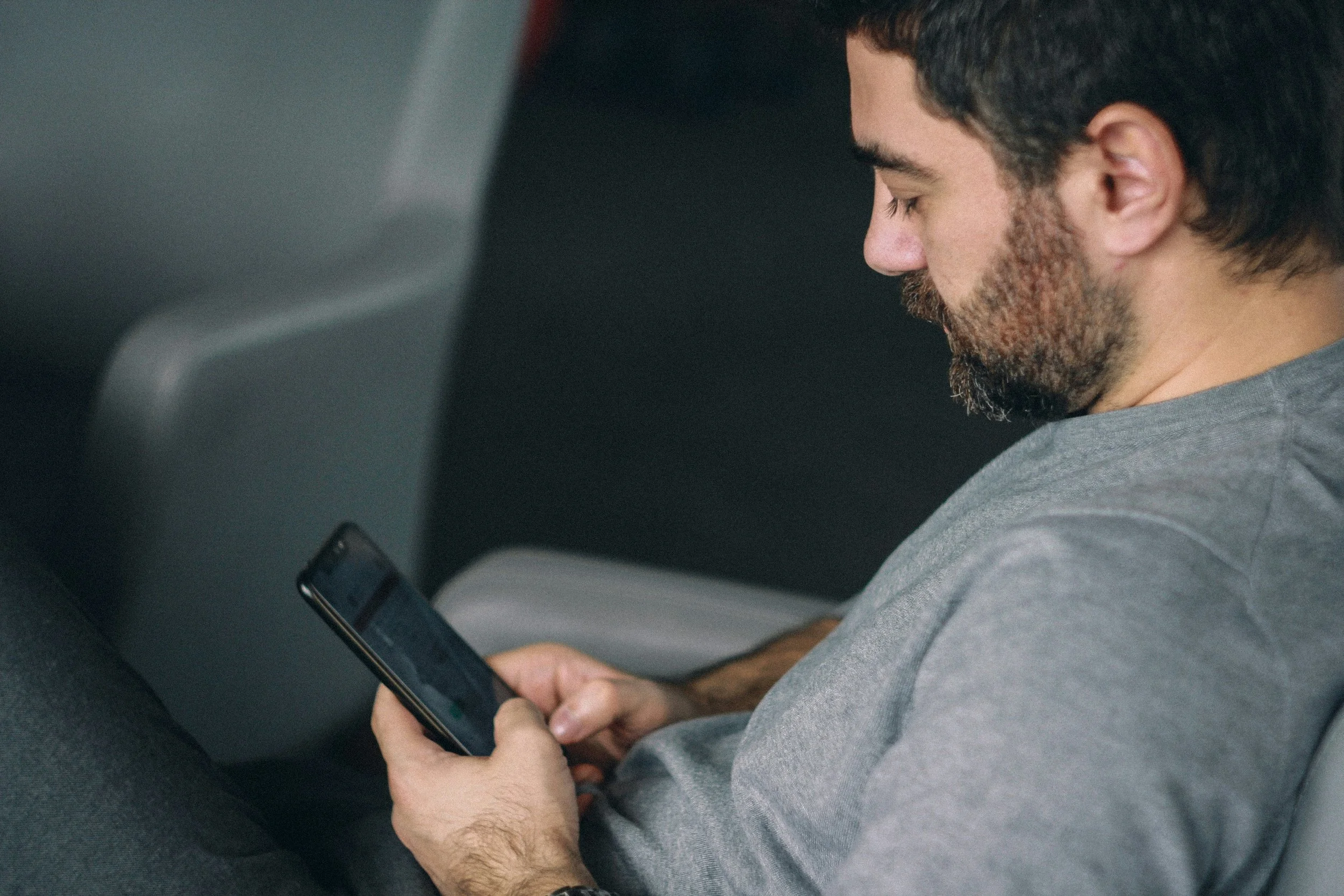 A bearded man in a grey sweatshirt looks at his phone while seated in a vehicle or public transport.
