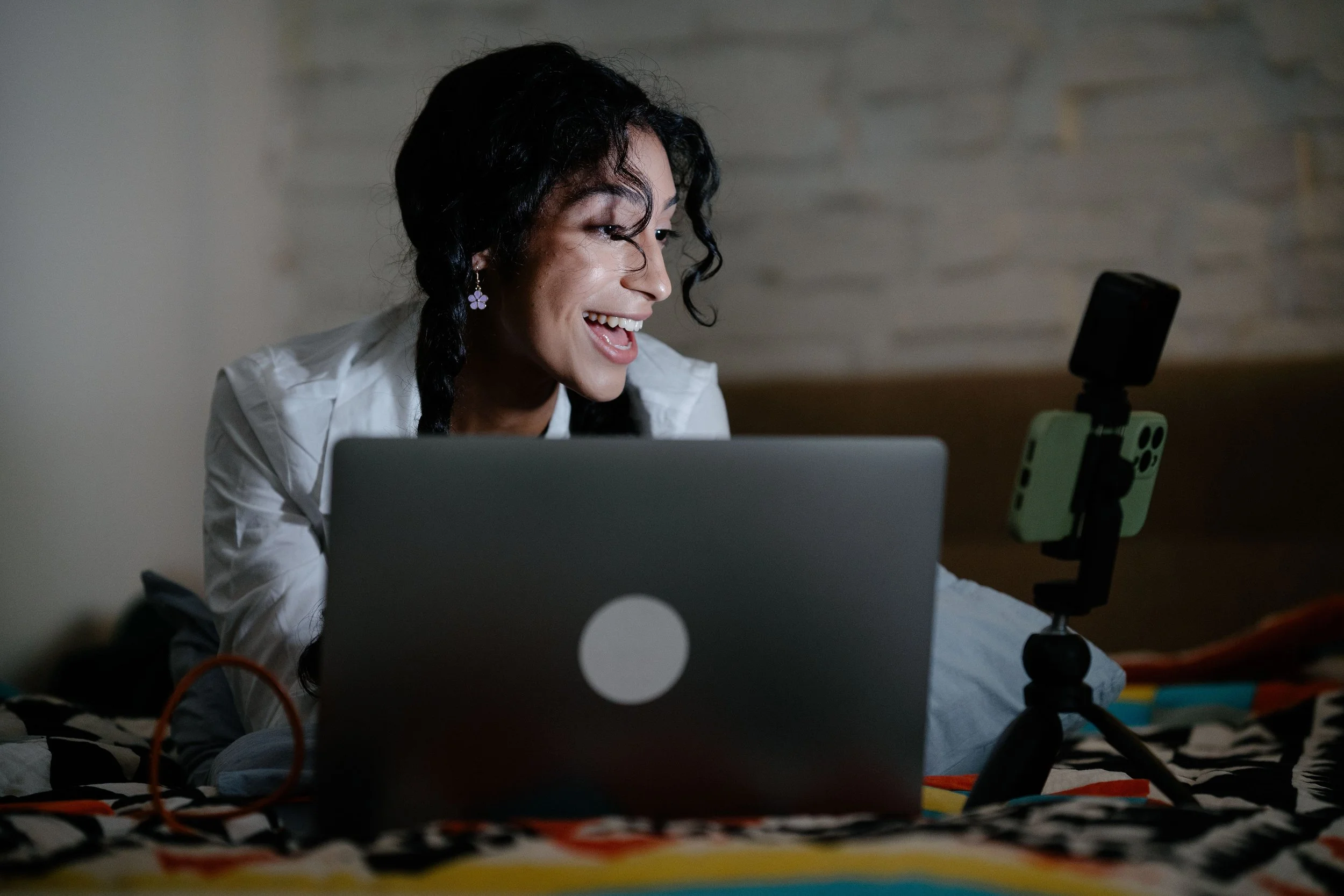A smiling woman lies on a bed using a laptop, with a phone on a small tripod beside her.