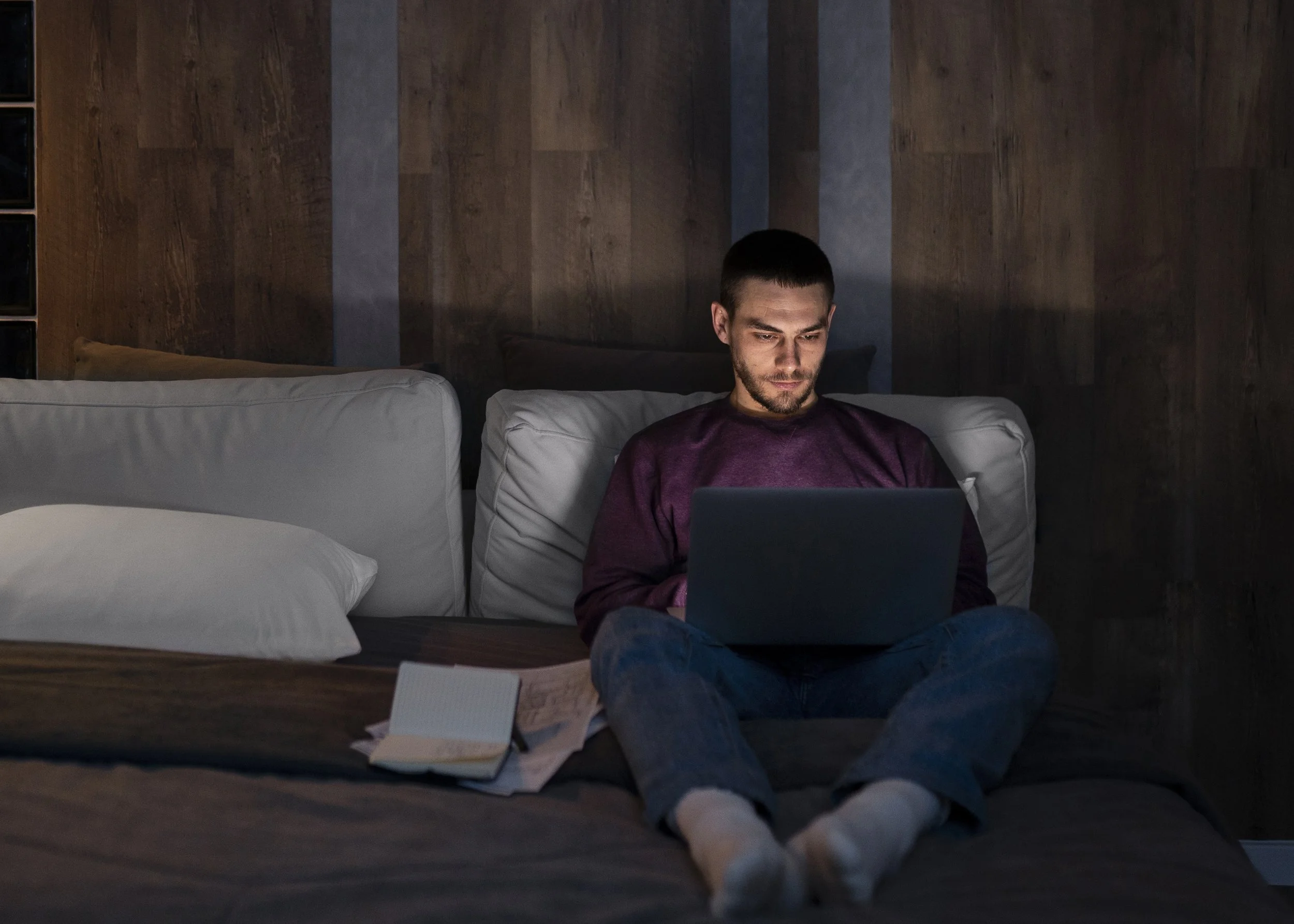 A man with short dark hair and a beard is sitting on a couch, looking at a laptop. He is wearing a maroon sweatshirt and blue jeans. The room has a dark wooden wall in the background and a beige cushion on the couch. There is a notebook, some papers, and a pen on the wooden surface in front of him, and a pair of white socks is visible in the foreground.