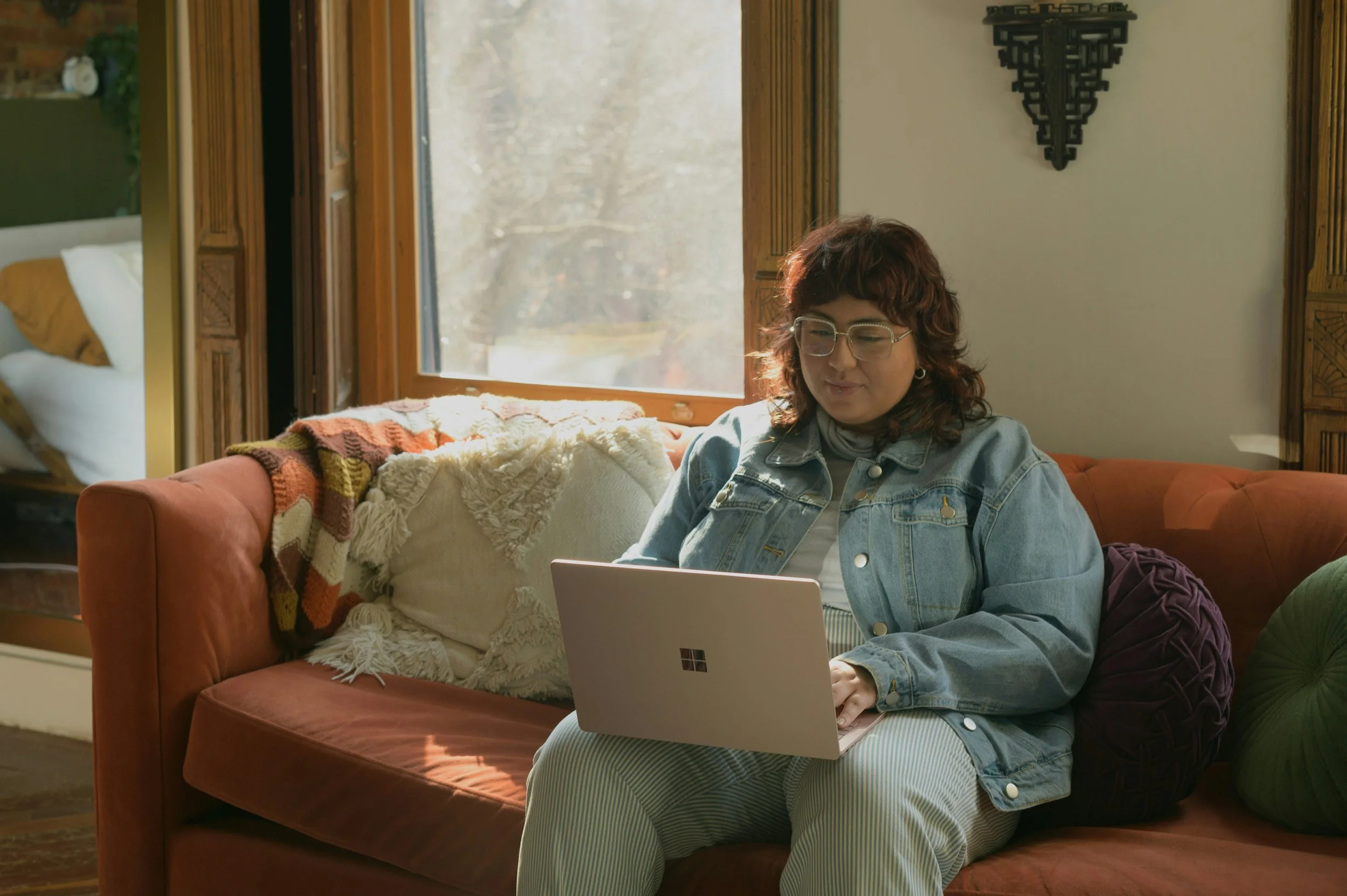 A woman with glasses and a denim jacket uses a Microsoft Surface laptop on a rust-coloured sofa.