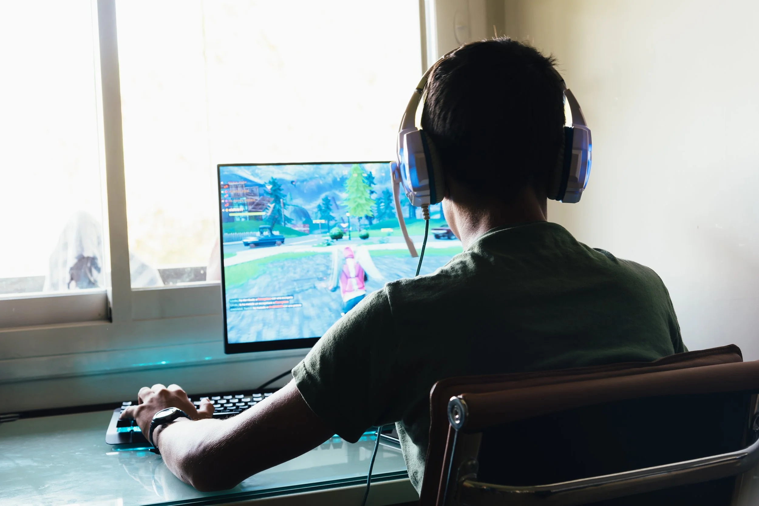 A young person wearing gaming headphones plays a video game at a desktop computer, viewed from behind.