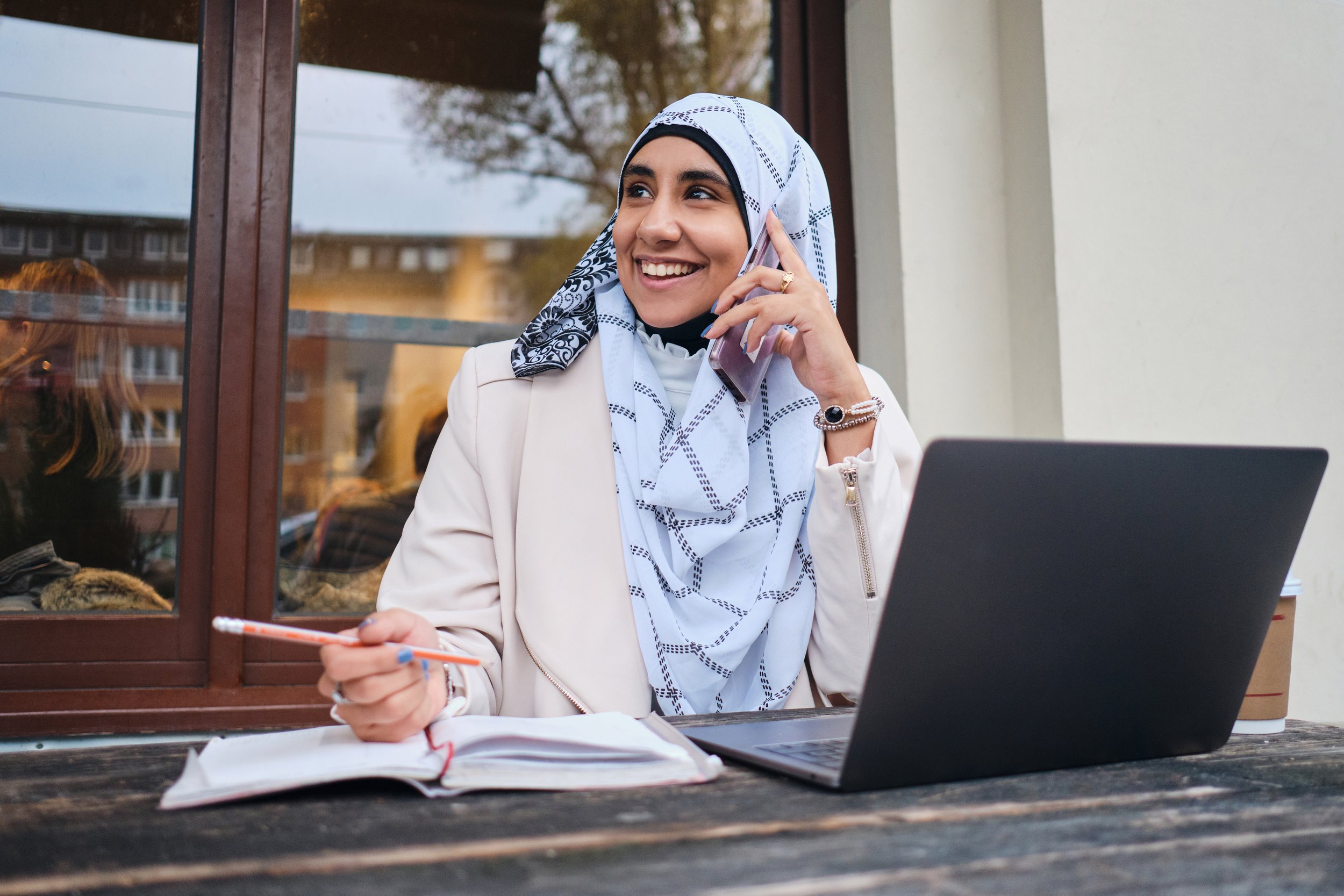 A woman wearing a white hijab, beige blazer, and jewelry is sitting outside, smiling on a phone call, with a laptop, planner, and notebook on the table in front of her.