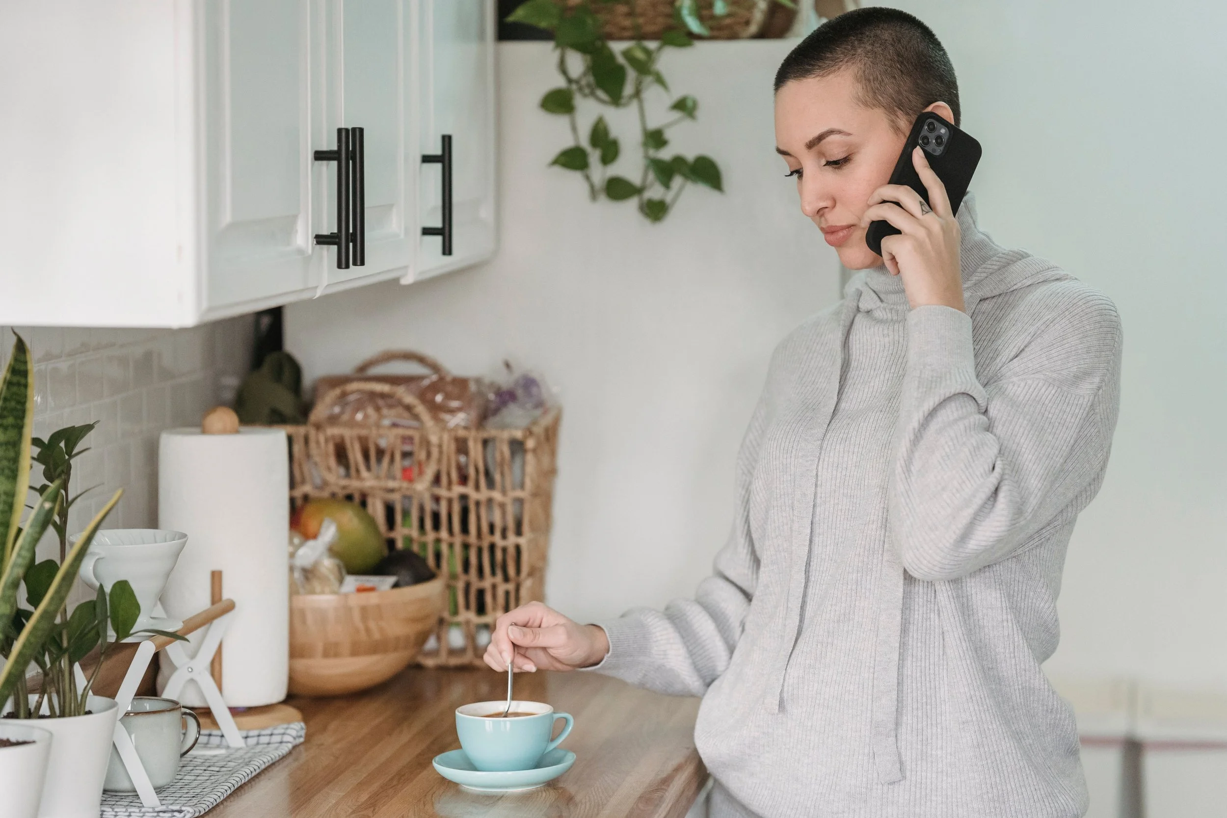 A person with a shaved head talks on the phone while stirring a cup of coffee in a kitchen.