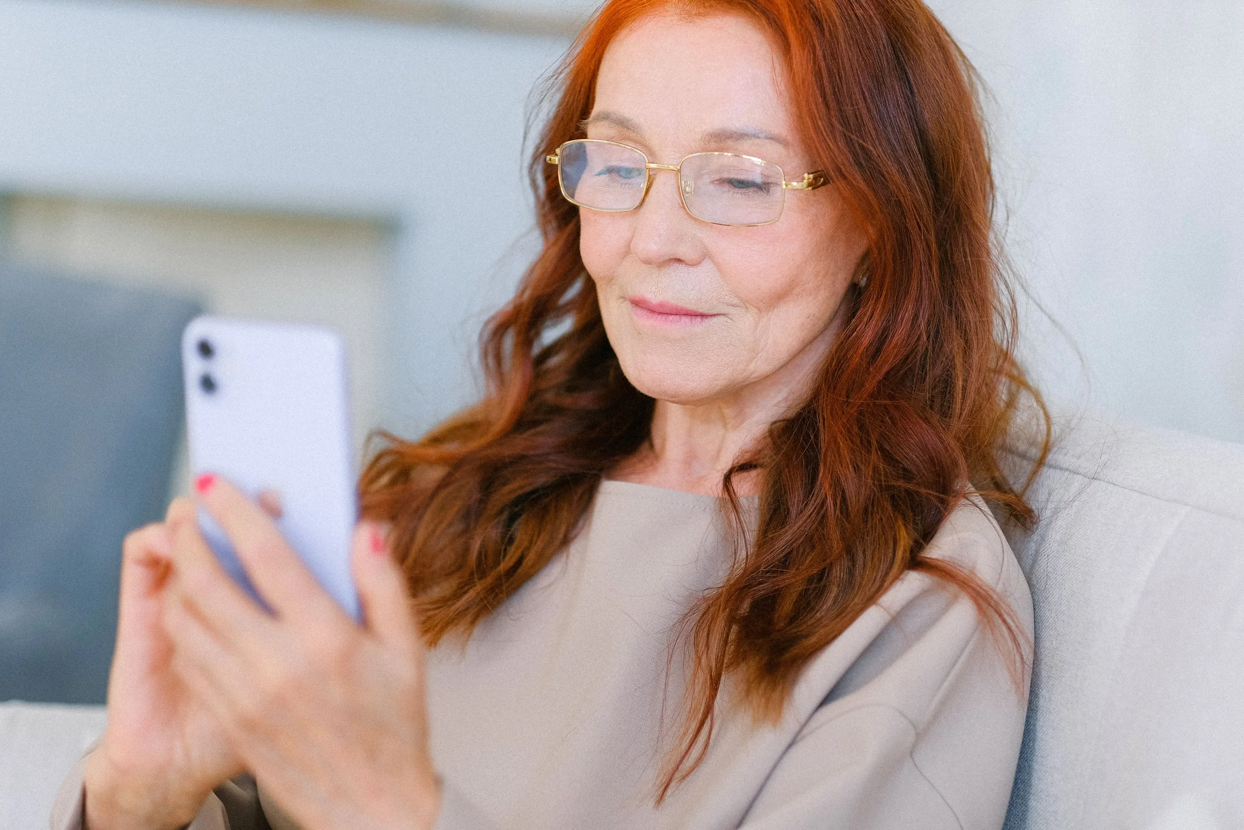An older woman with red hair and gold-framed glasses looks at her phone while seated on a sofa.