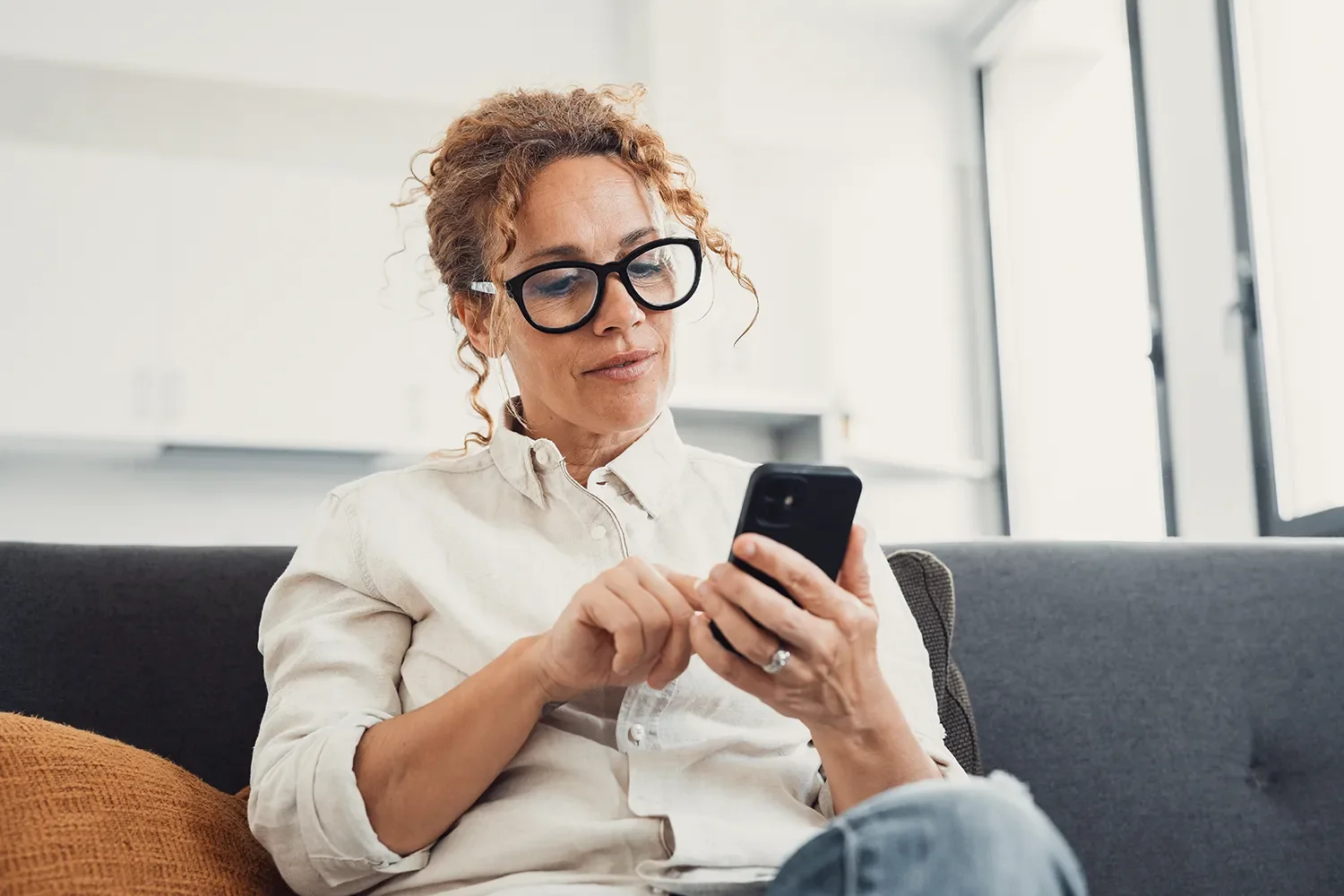 Woman with curly hair and glasses sitting on a black couch, using her smartphone in a bright room with large windows.