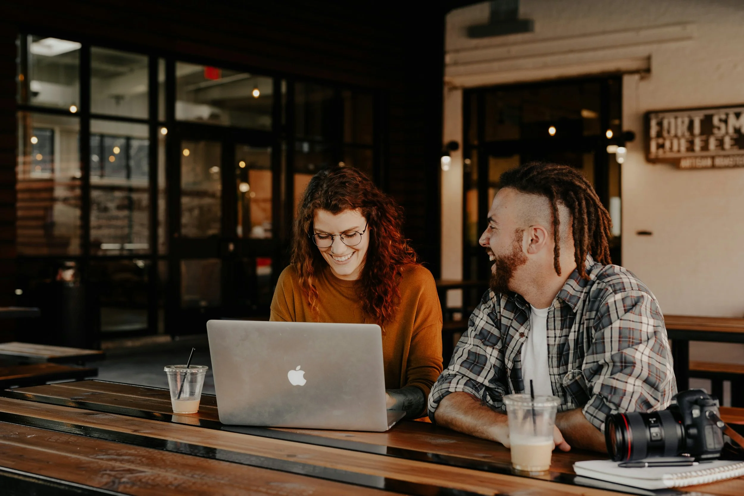 Two people, a woman with curly red hair and glasses and a man with dreadlocks and a beard, sitting at a wooden table in a coffee shop, looking at a laptop together. There are drinks on the table, one with a straw and a lid, and a camera near the edge of the table. The background shows a modern cafe interior with large windows and warm lighting.