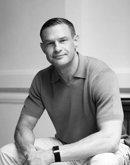 A black and white photo of a man sitting casually, wearing a short-sleeved polo shirt, with short hair, in an indoor setting.