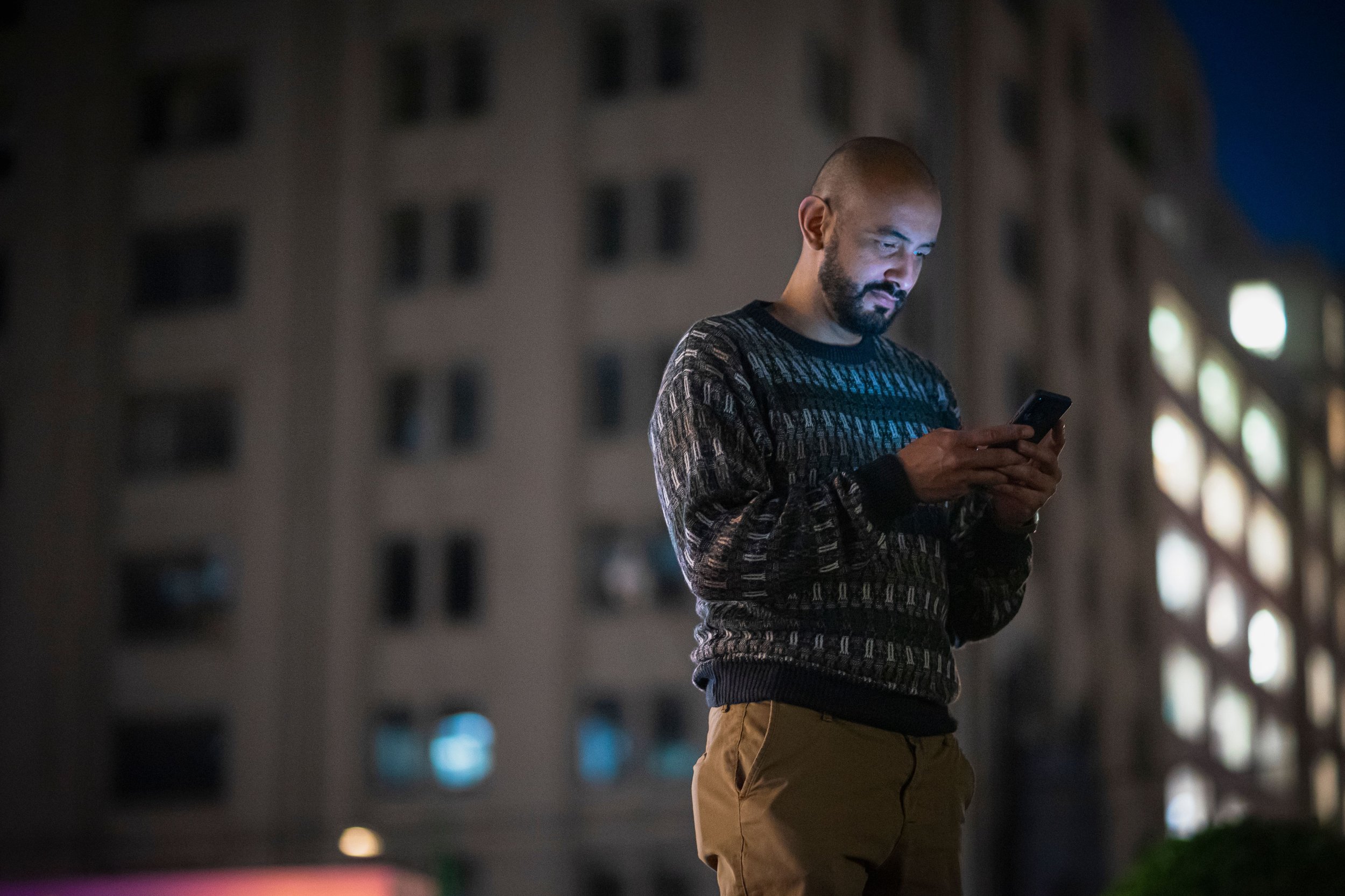 A man stands in front of a building at night, looking at his phone with city lights in the background.