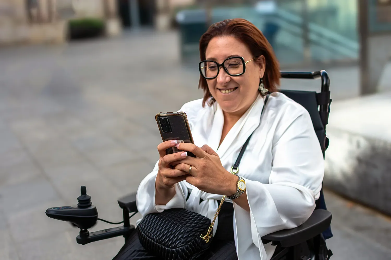 A woman with short red hair and glasses sitting in a wheelchair outdoors, smiling as she looks at her smartphone.