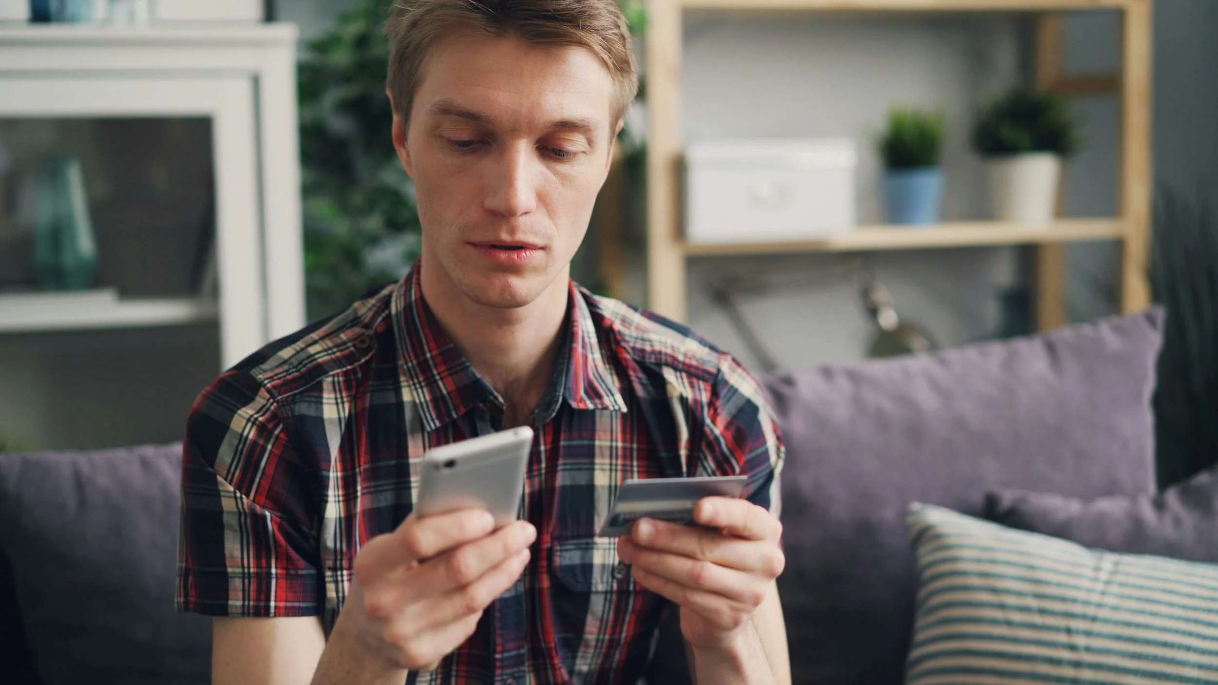 A man sits on a couch, holding a credit card in one hand and a cell phone in the other.
