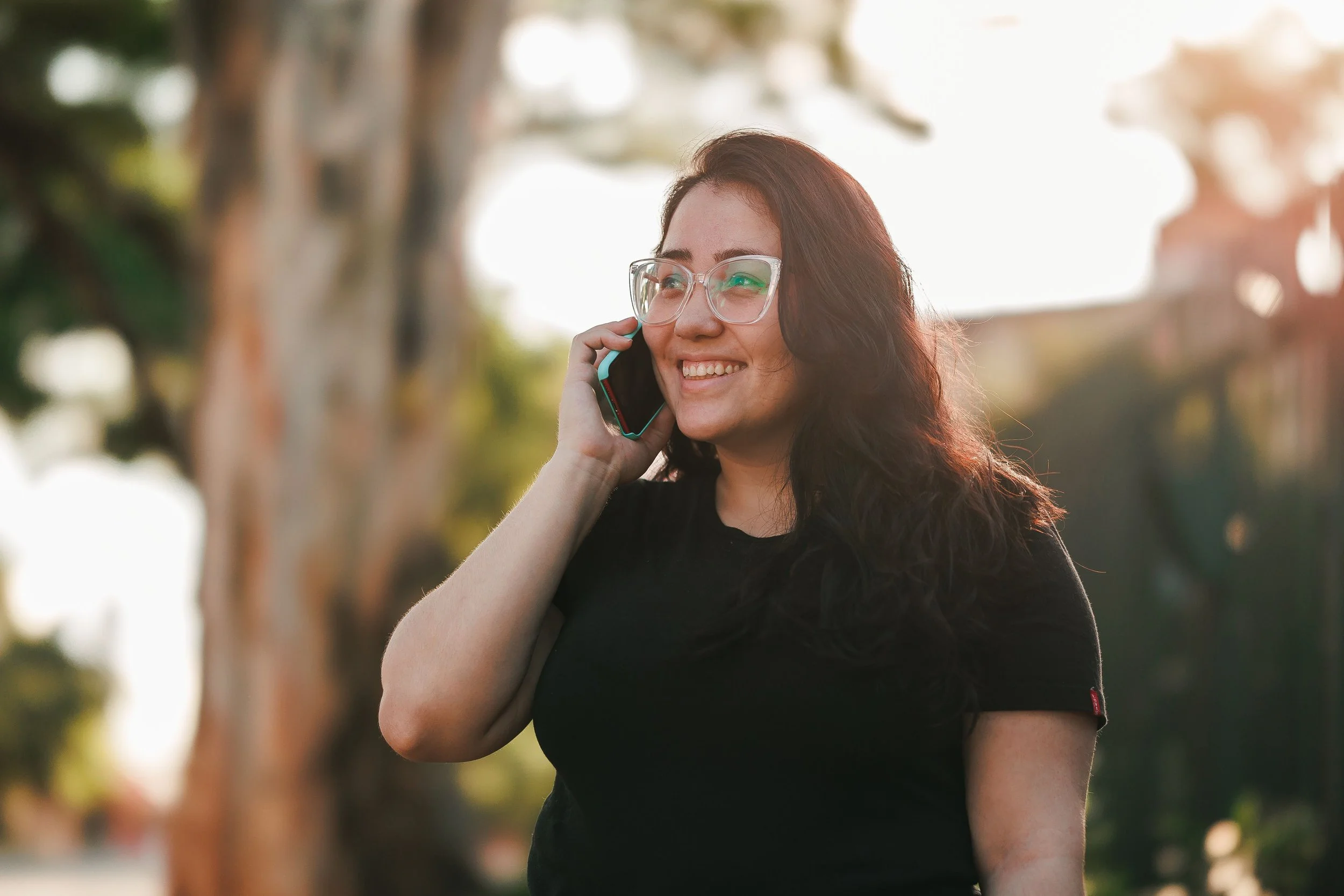 A smiling woman with glasses and long dark hair talks on a phone outdoors in warm sunlight.