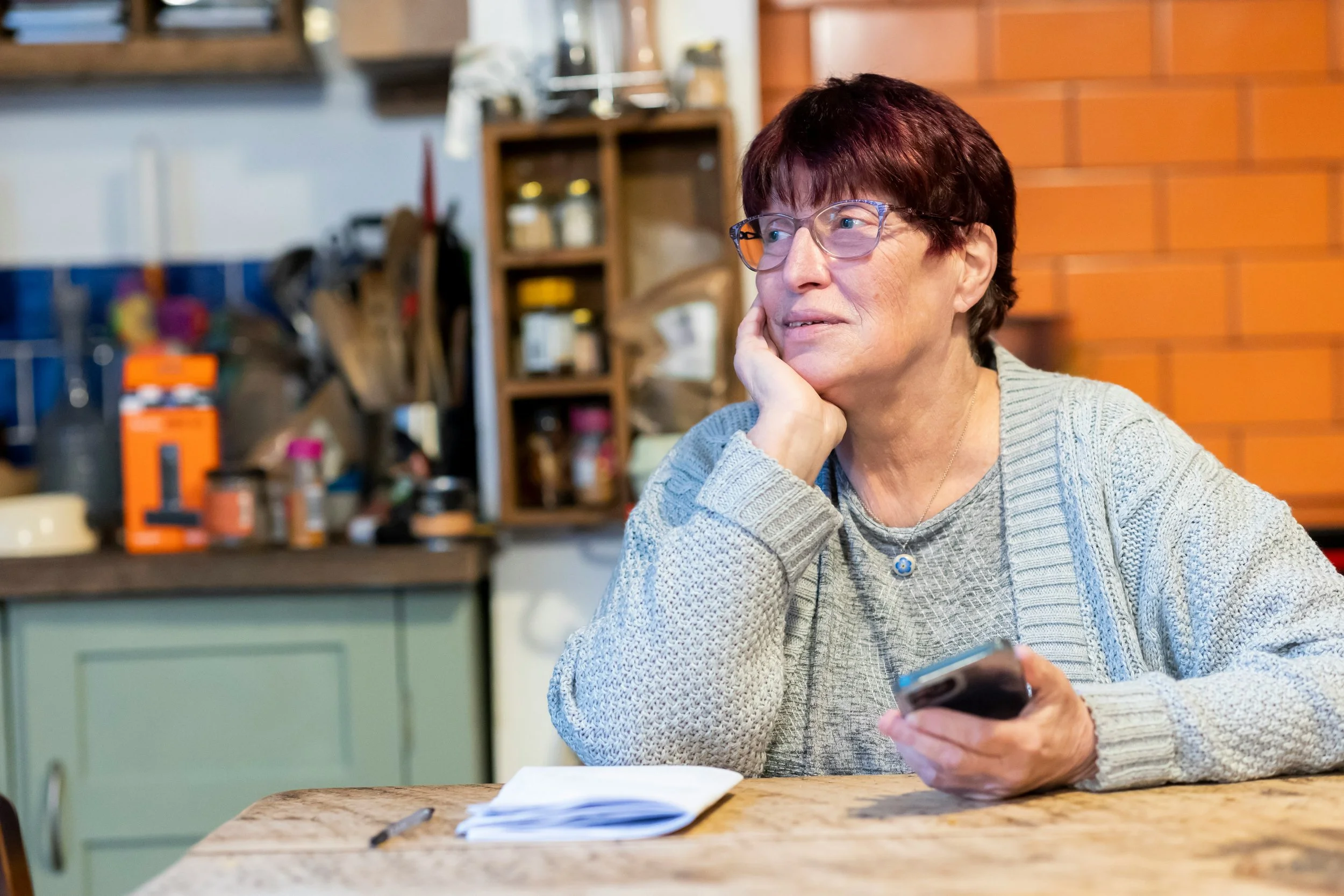 An older woman with glasses and a blue cardigan sits at a kitchen table holding a phone, looking thoughtfully into the distance.