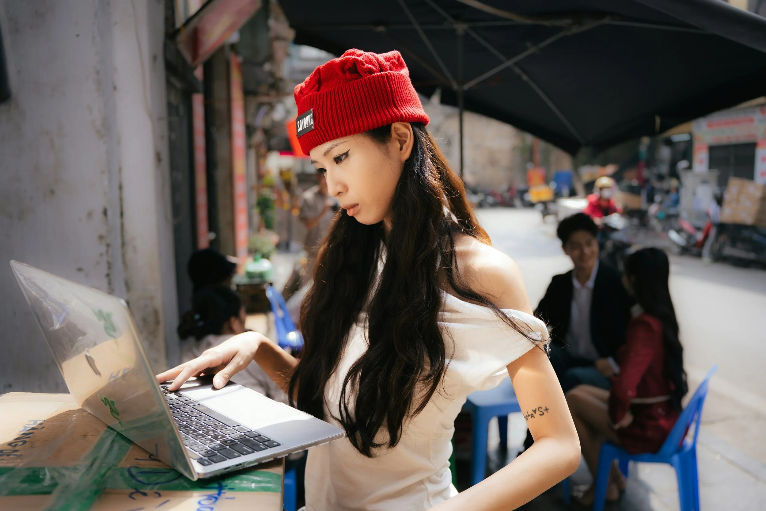A woman in a red beanie and white top leans over a laptop on a street-side table in a busy urban setting.