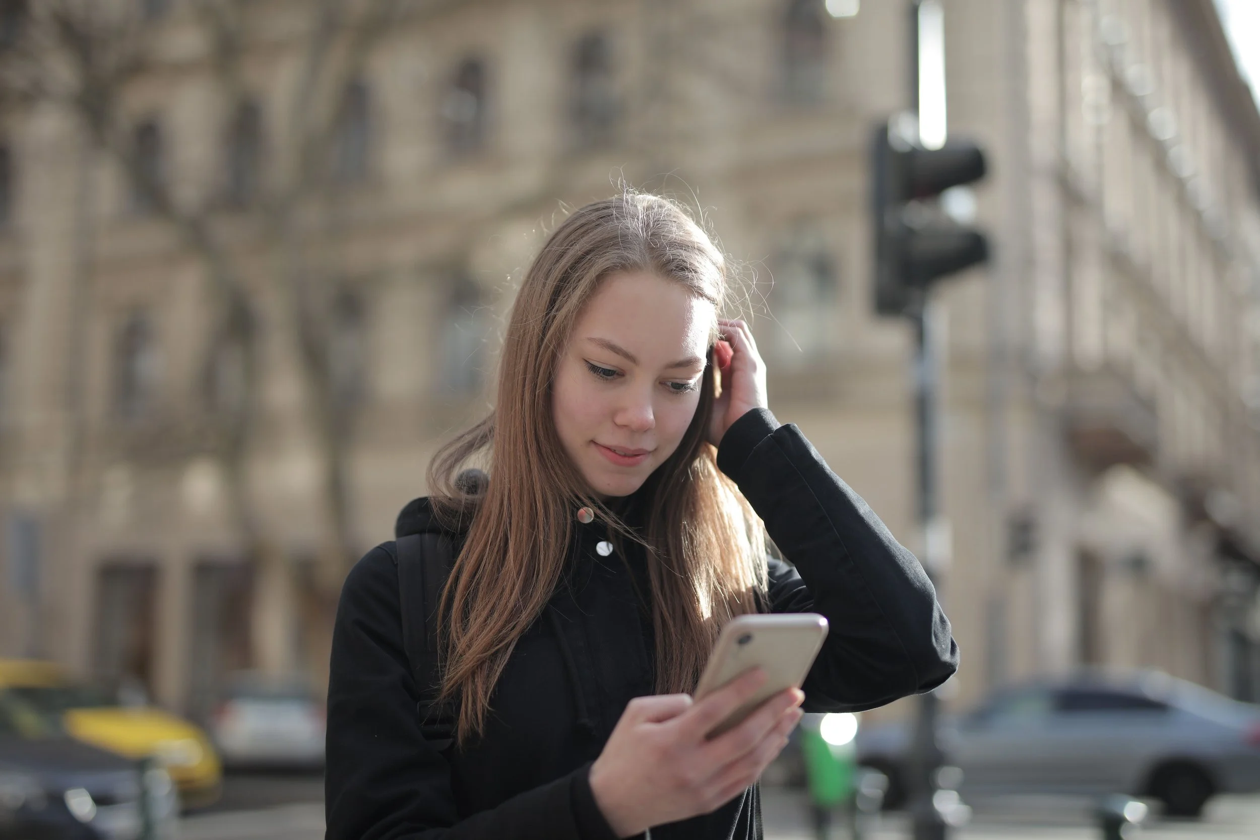 A young woman with long hair looks at her phone on a city street, with buildings and a traffic light behind her.