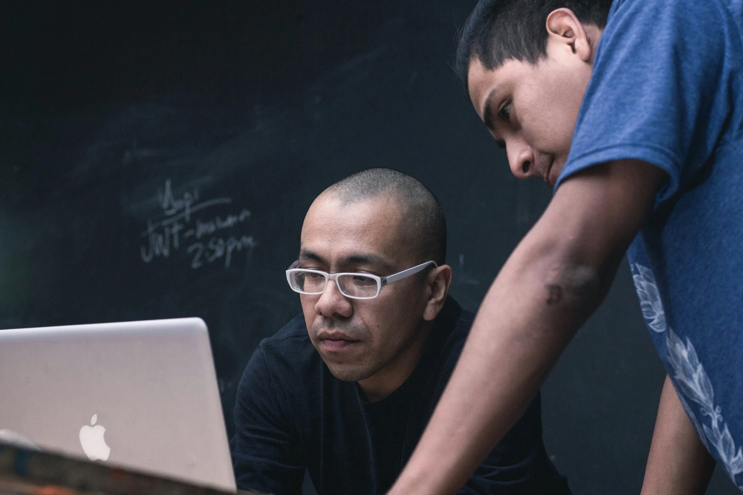 Two men are working together at a computer. One man, with short dark hair and glasses, is sitting in front of the laptop, while the other, with short dark hair, is standing over him, leaning in to look at the screen. They are in a room with a blackboard or chalkboard in the background.