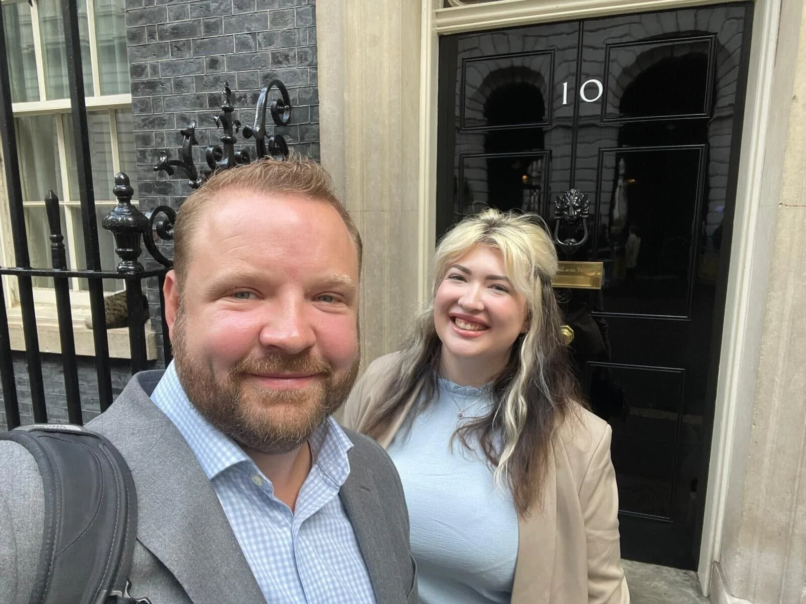 A man with a beard and a woman with blonde hair standing in front of a black door with the number 10 on it, smiling at the camera.