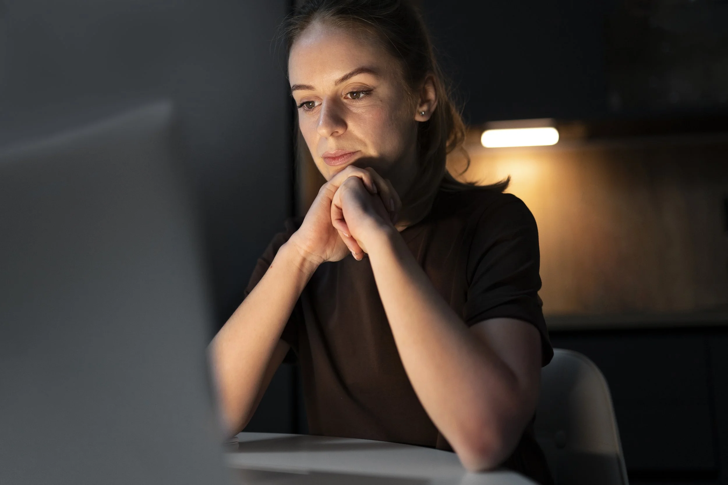 A woman with brown hair and a thoughtful expression, resting her chin on her hand, sits at a desk in a dimly lit room.