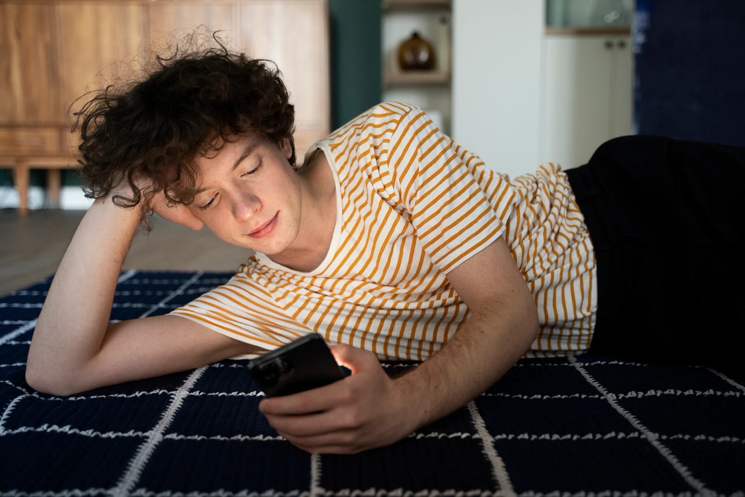 A young person with curly hair lies on a rug, looking at their phone in a bright living room.