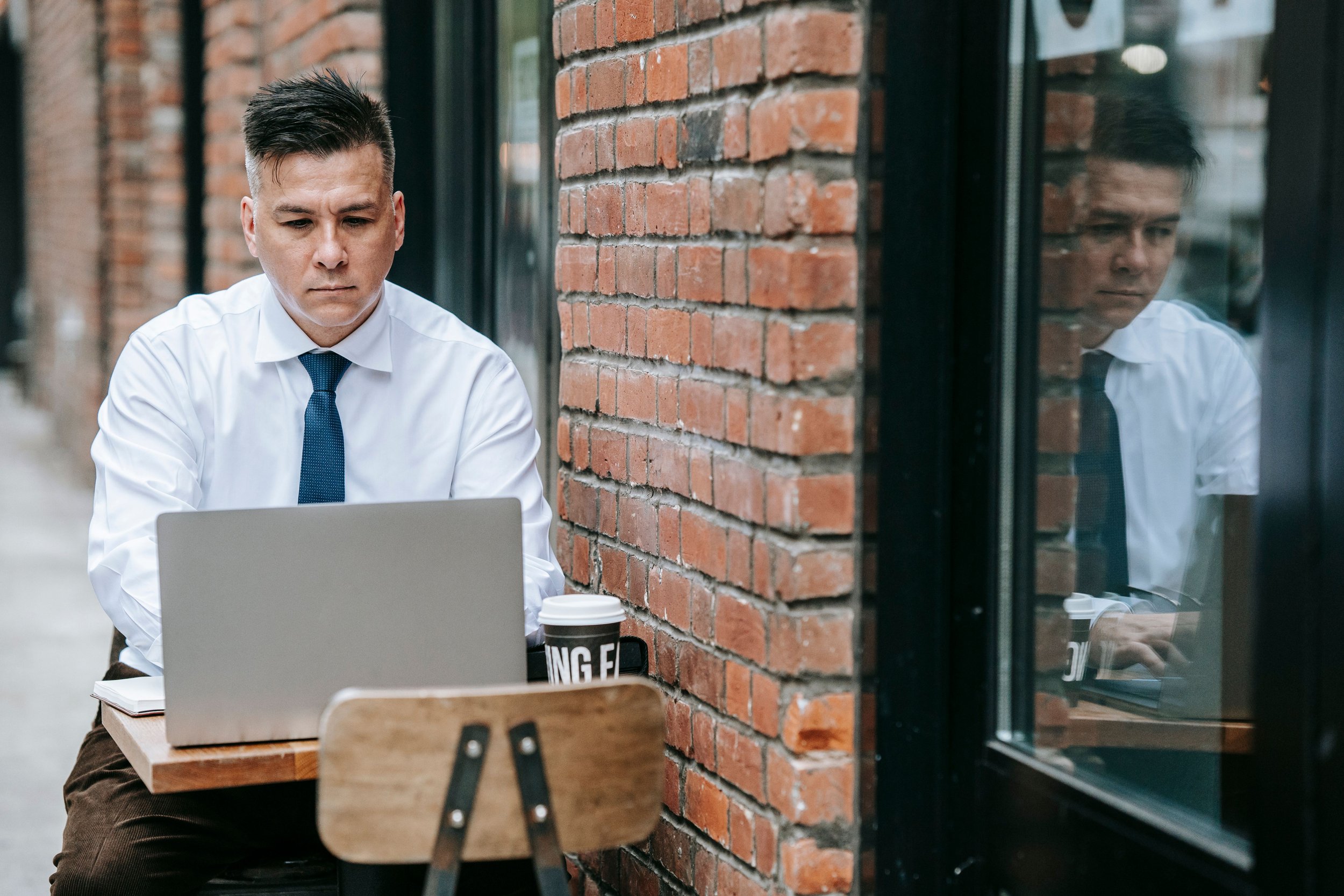 A man in a white shirt and blue tie works on a laptop at an outdoor café table beside a brick wall.