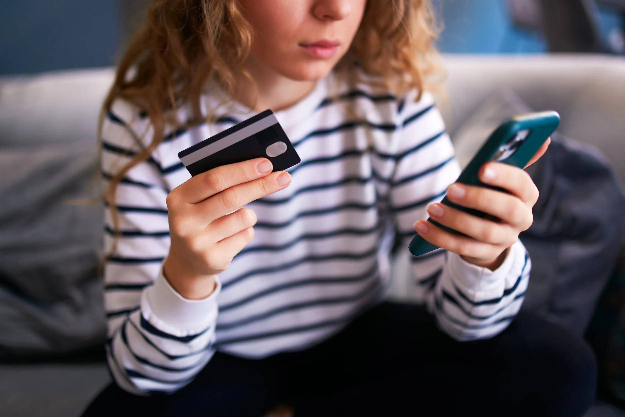 A woman holds a bank card and phone while seated on a sofa.