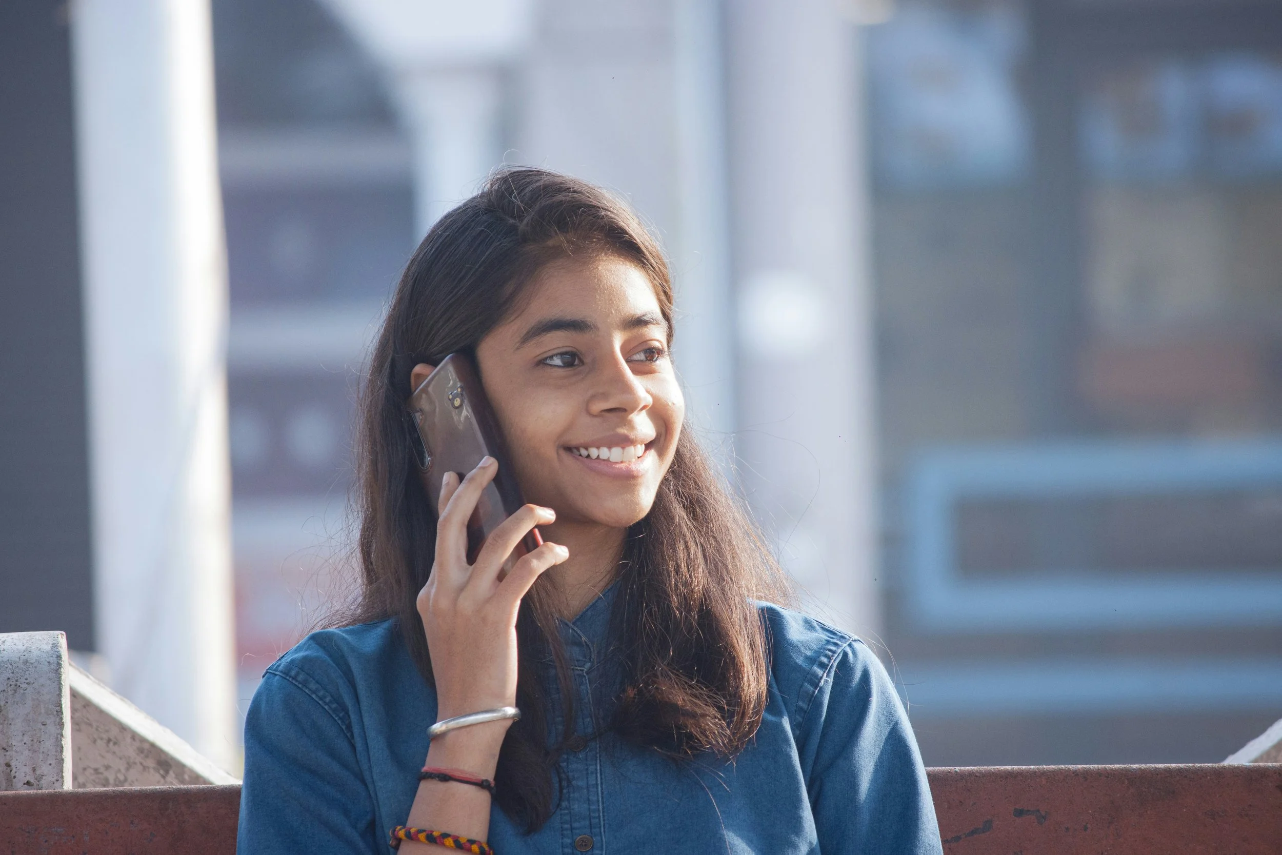A young girl with long dark hair smiles while talking on a phone outdoors.
