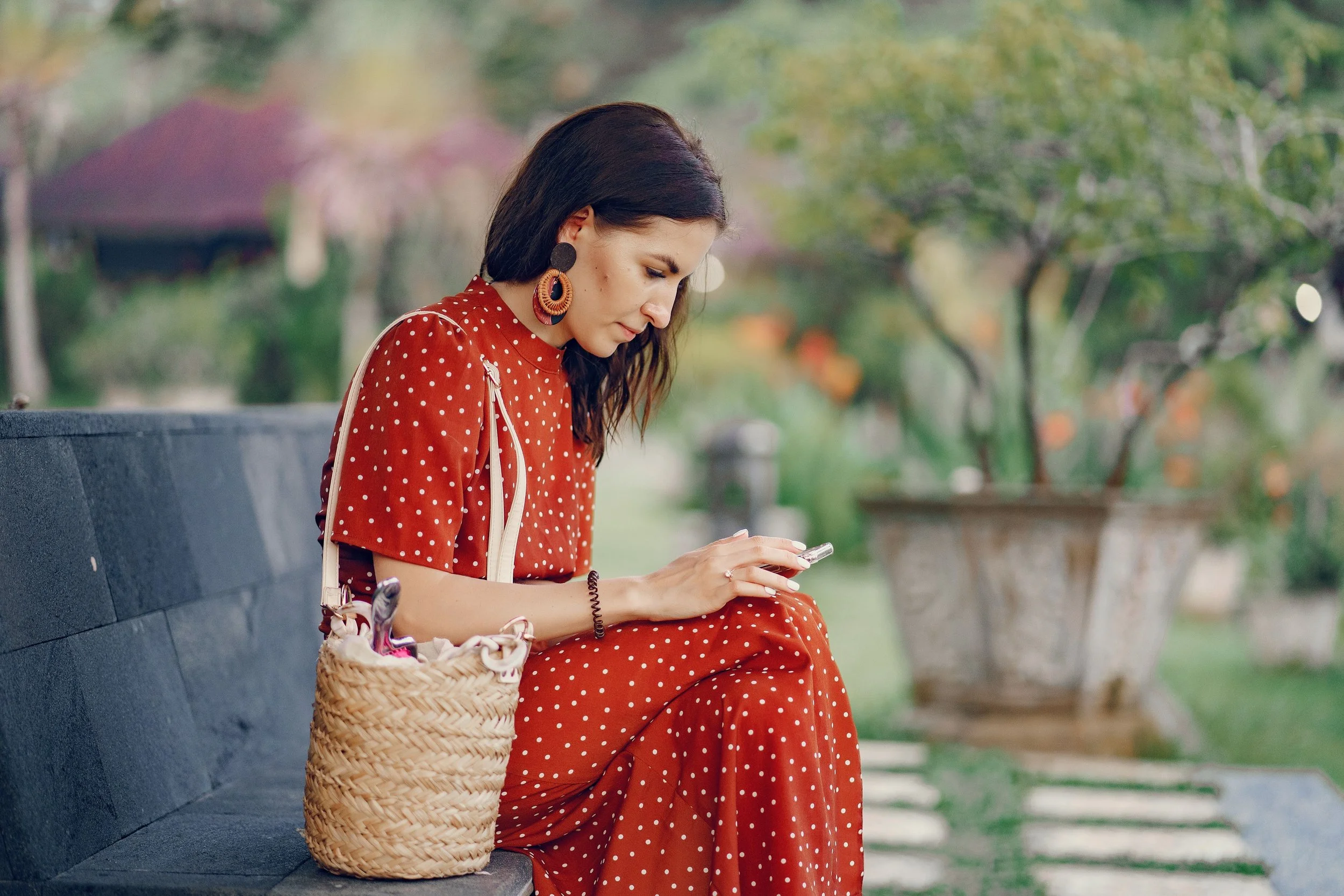 A woman in a red polka dot dress sits on a bench outdoors, looking at her phone, with a wicker bag beside her.