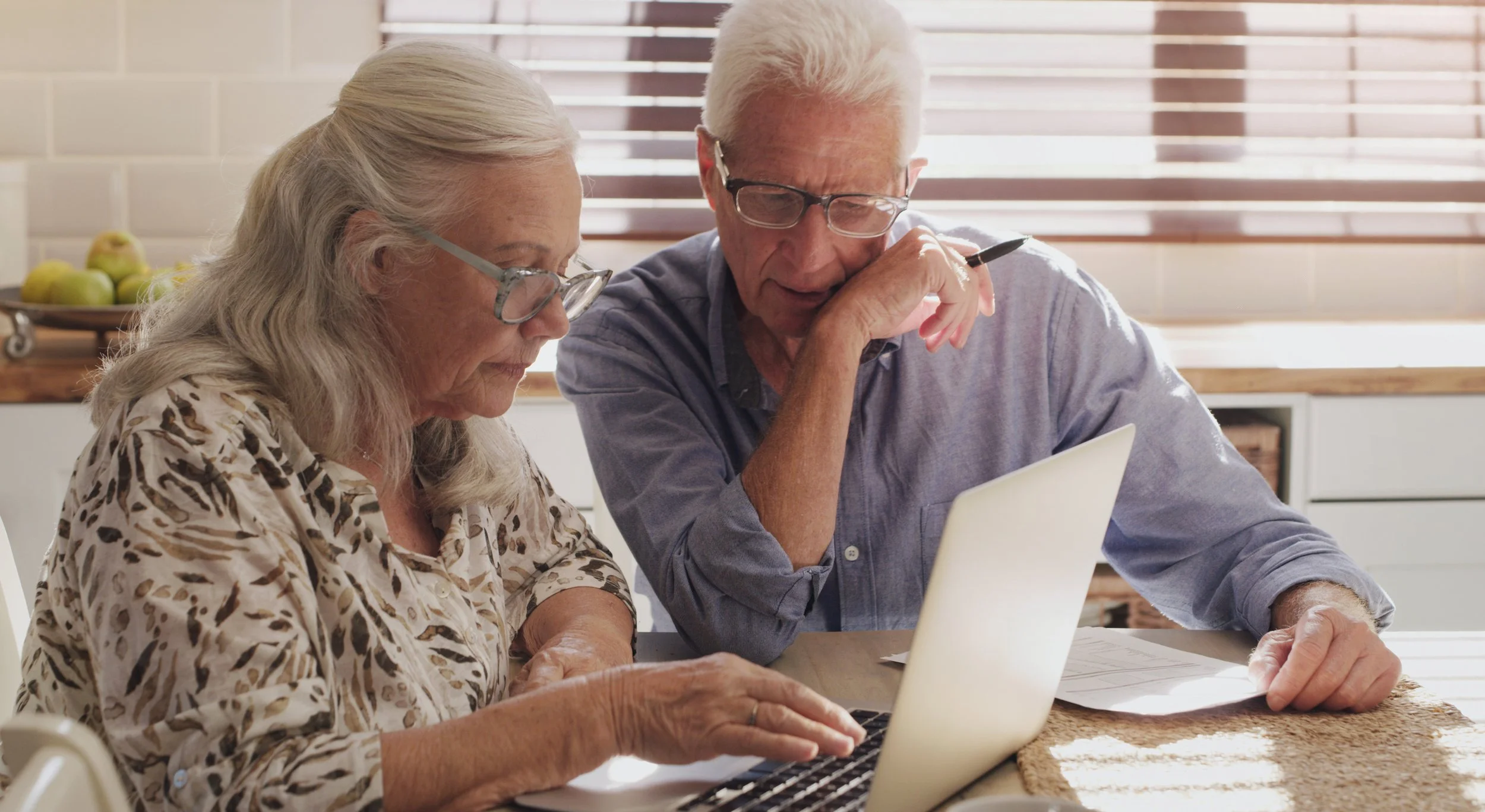 An elderly man and woman sitting at a table using a laptop together in a bright kitchen.