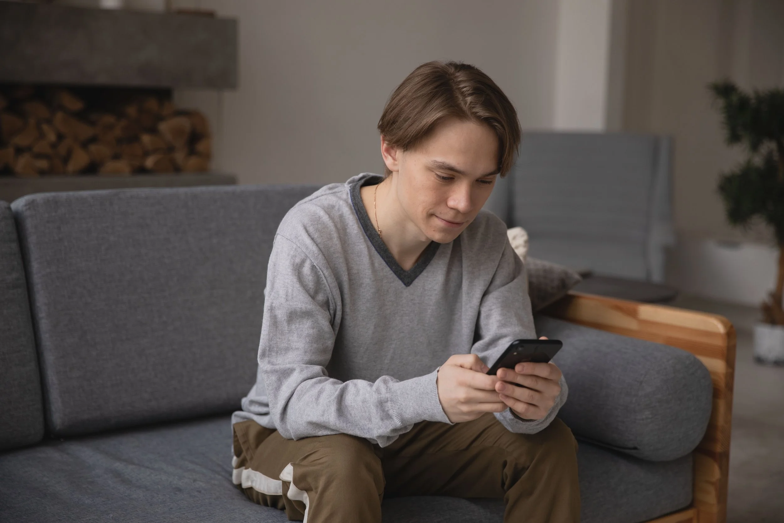 A young man in a grey jumper sits on a sofa looking at his phone in a modern living room.