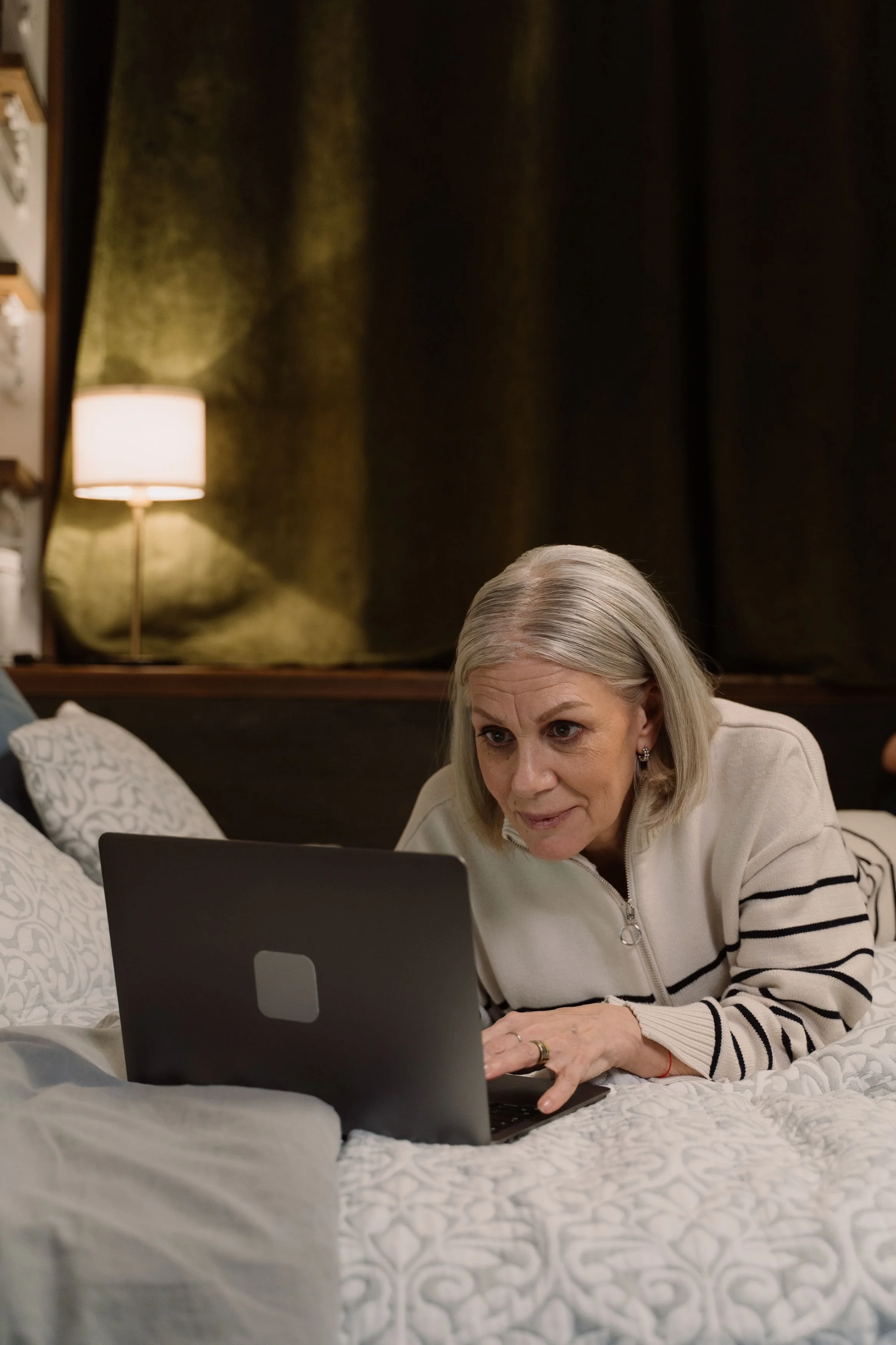An older woman with silver hair lies on a bed, looking intently at a laptop screen in a dimly lit room.