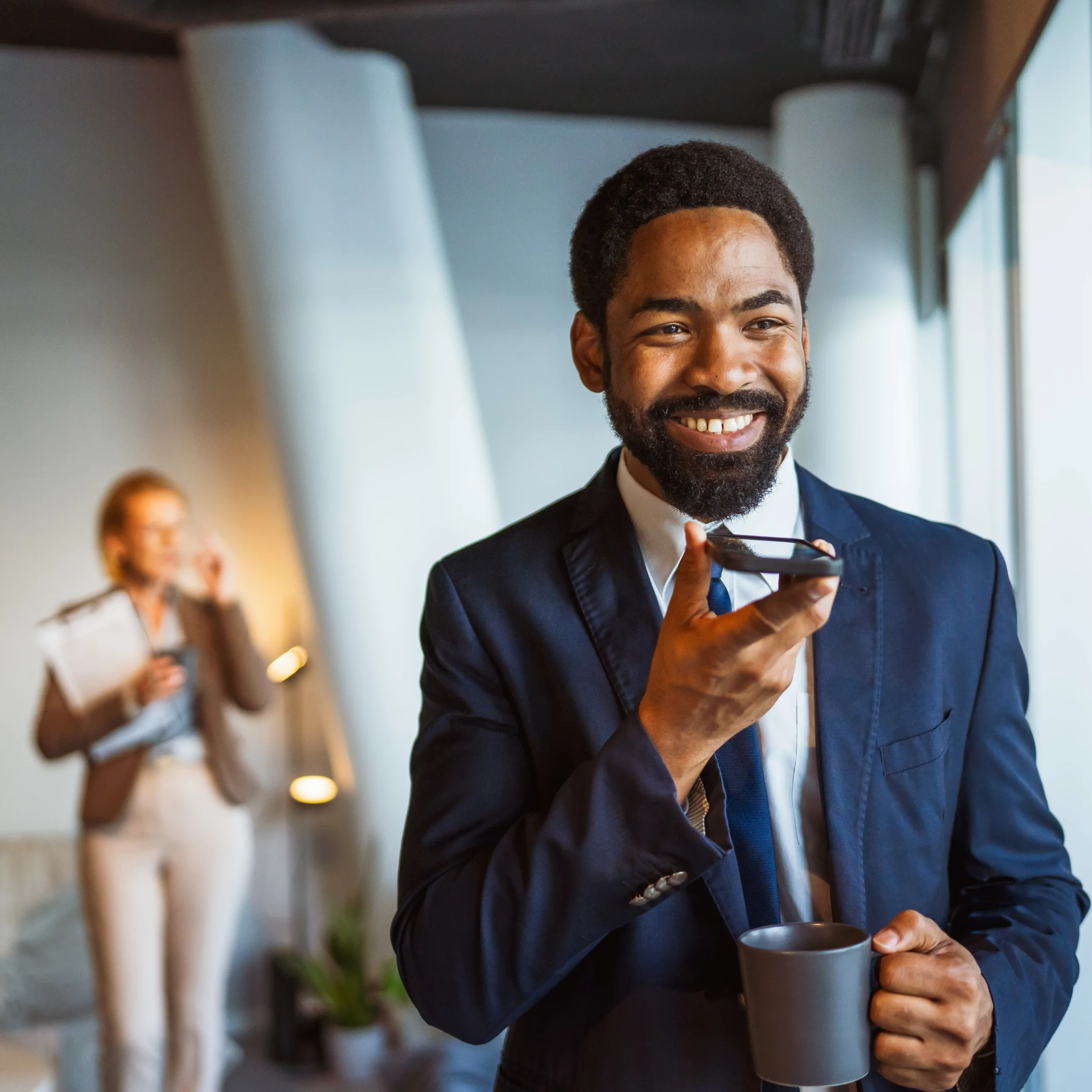 Smiling man in a formal suit holding a gray coffee mug, blurred background with people and windows in a bright indoor setting.