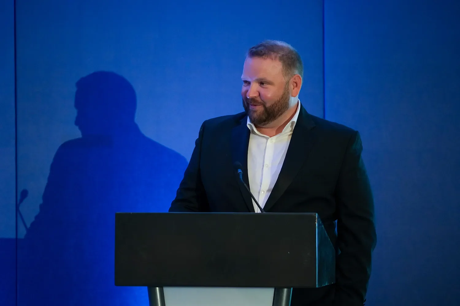 Our CEO Rory Innes with a beard and suit stands at a podium during a presentation or speech, with a shadow of himself cast on the blue wall behind him.