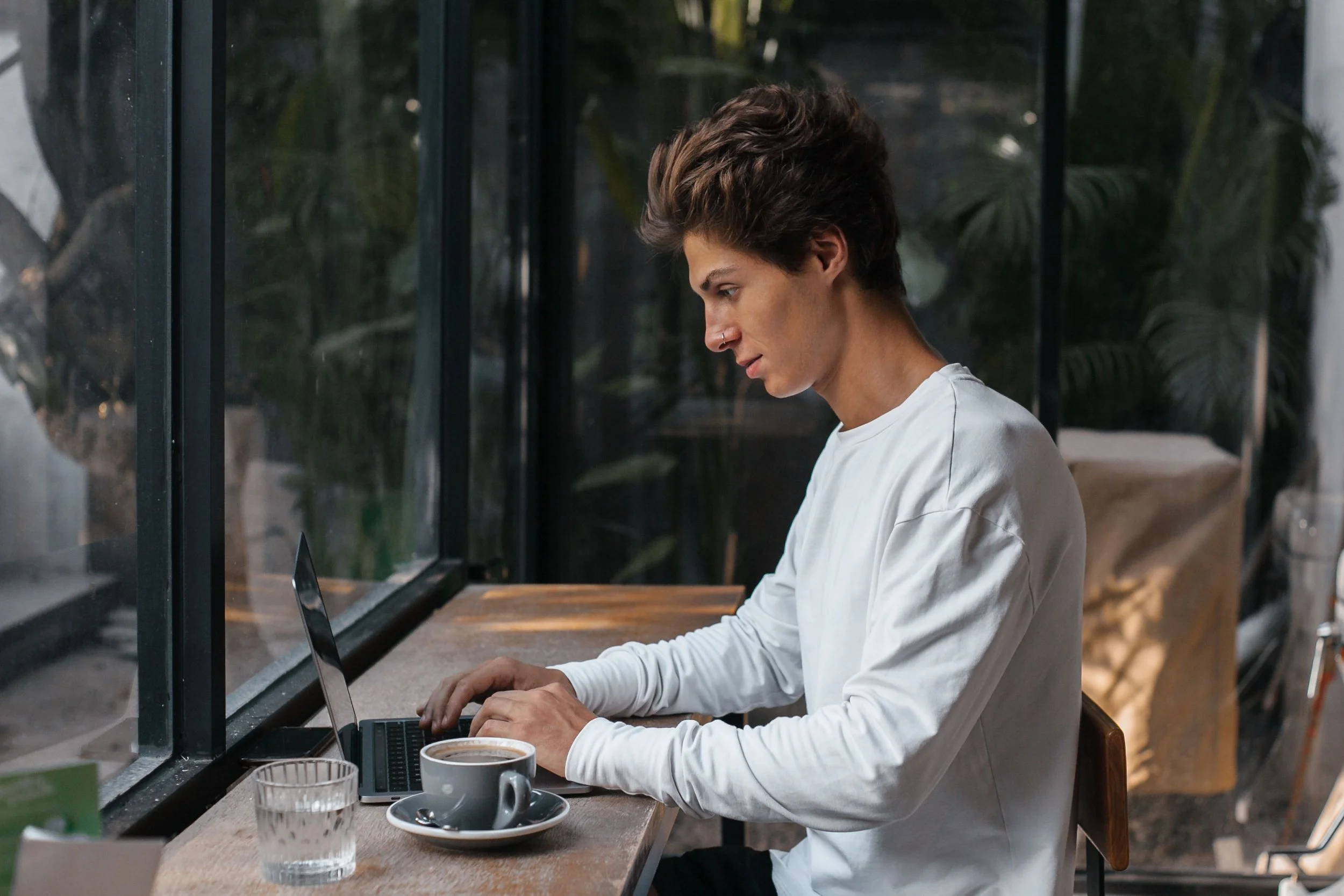 A young person in a white sweatshirt types on a laptop at a café table, with a coffee cup beside them.