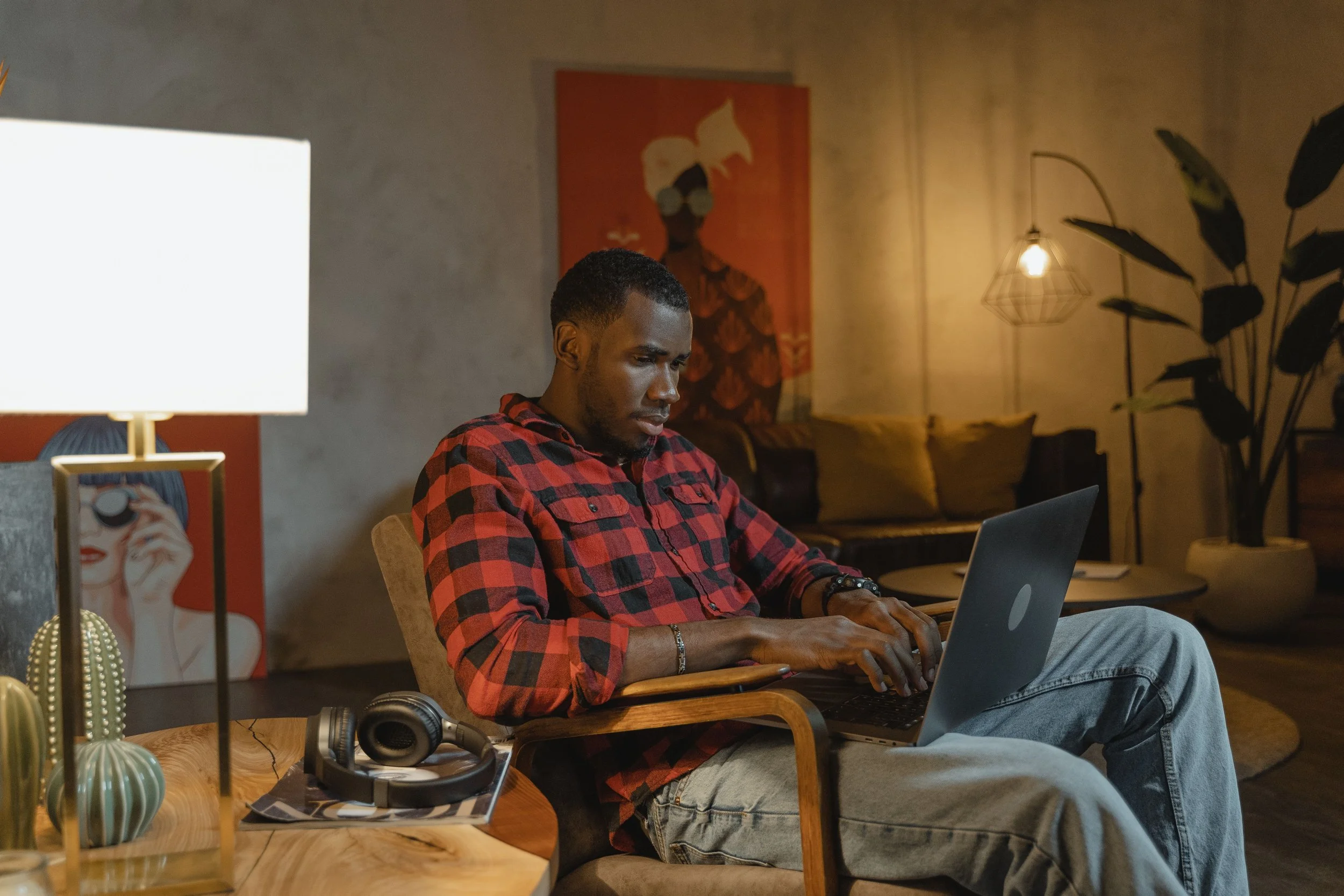 A man in a red checked shirt types on a laptop in a warmly lit, stylishly decorated living room.