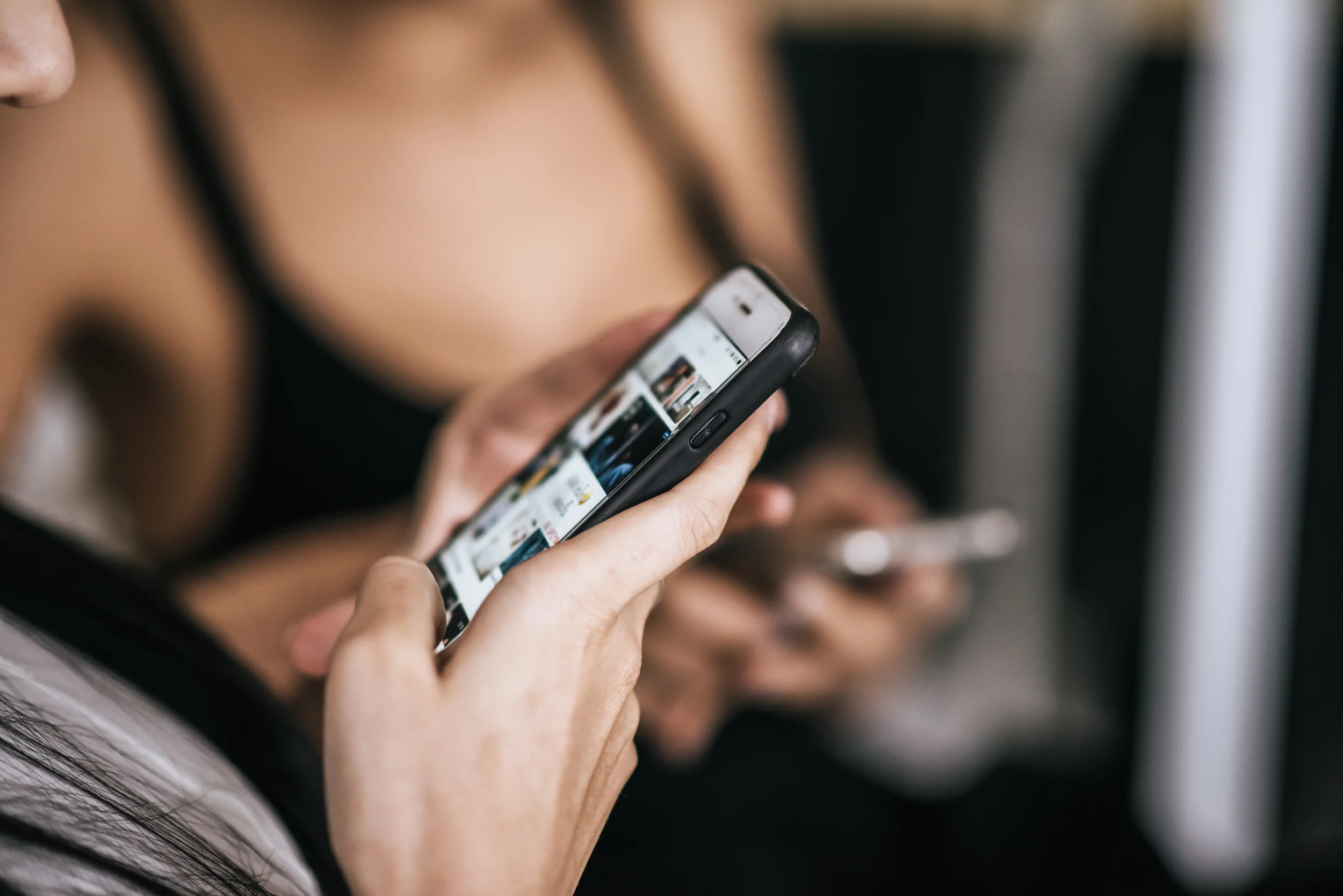 A close-up of a hand scrolling through a social media feed on a phone, with a second person holding a phone in the background.