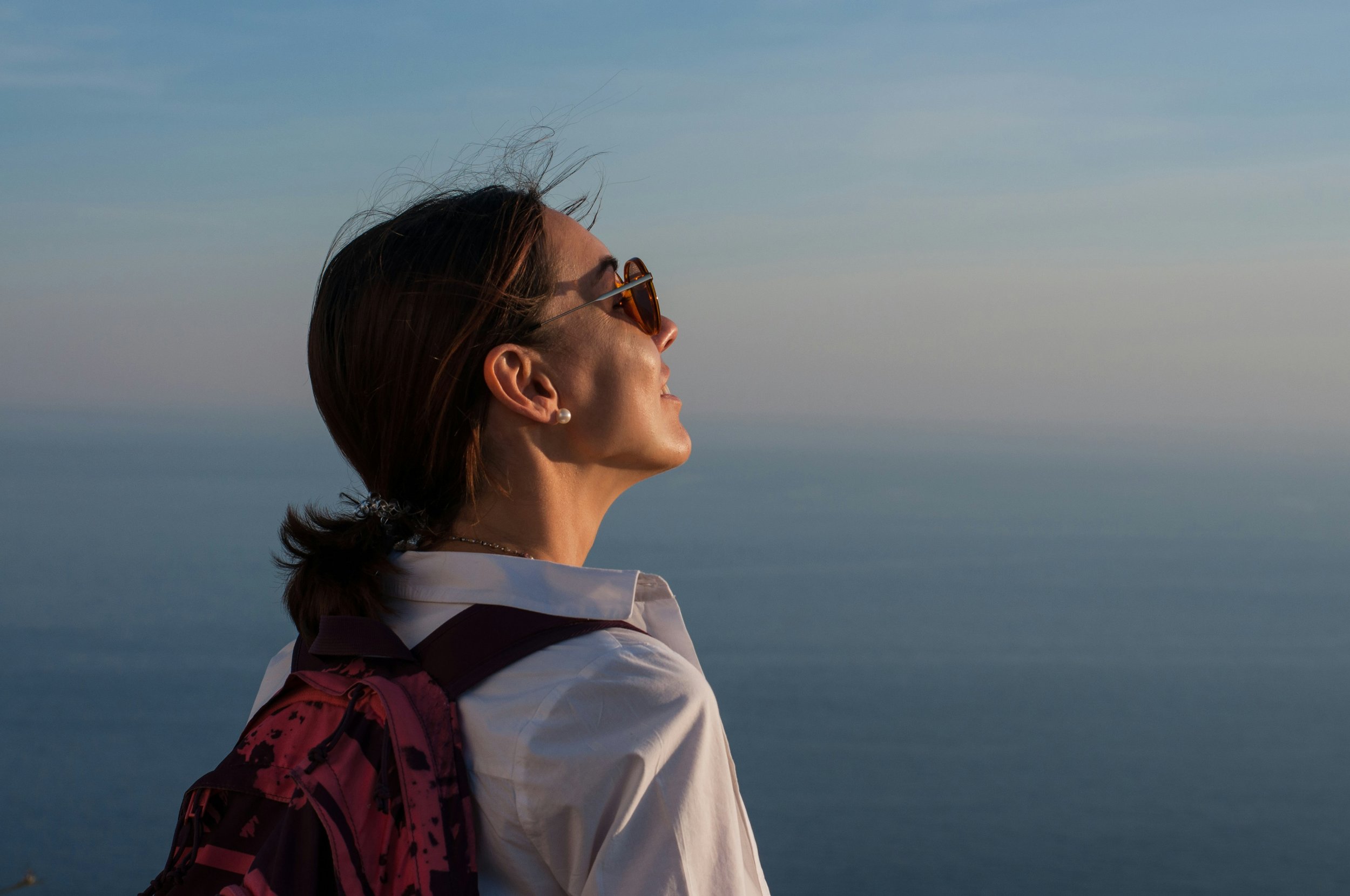 A woman wearing sunglasses and a backpack looks out over a calm, blue body of water under a clear sky.