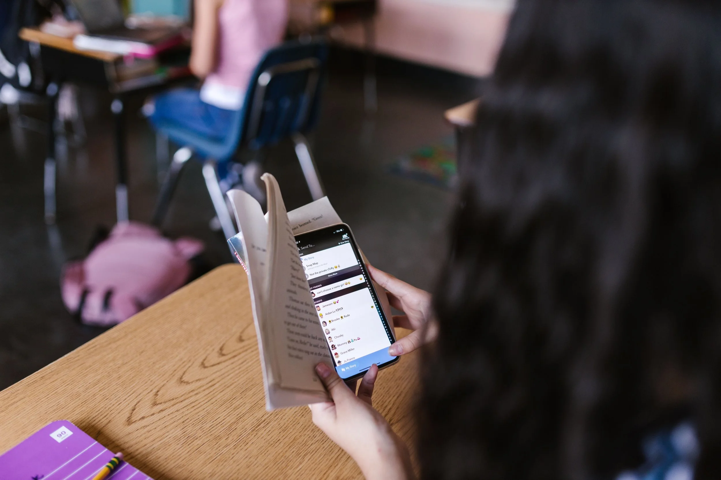 A pupil hides a phone showing Snapchat behind a book at a school desk during class.