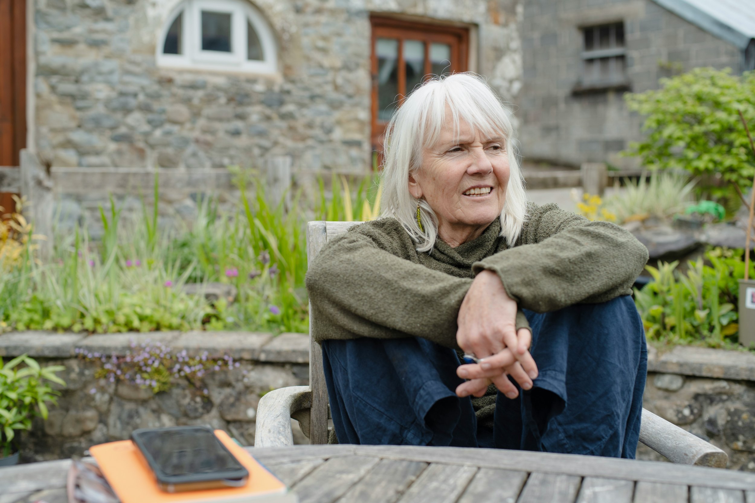 An older woman sits on a wooden chair in front of a stone building, smiling and enjoying the outdoors.